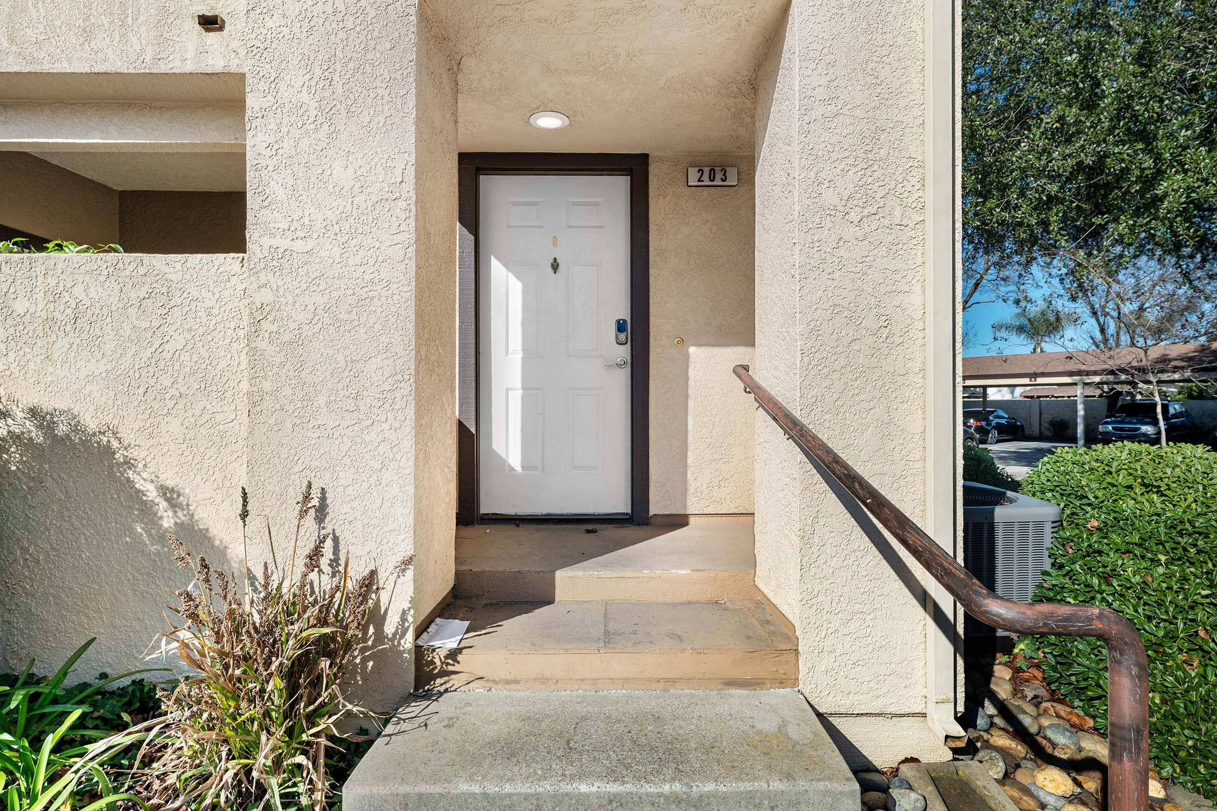 Entrance to a unit with a white door labeled "203." The area features textured beige walls, a small set of steps, and landscaping with green plants. A light fixture is mounted above the door, and there are railing and stone details visible. Sunlight illuminates the scene, indicating a clear day.
