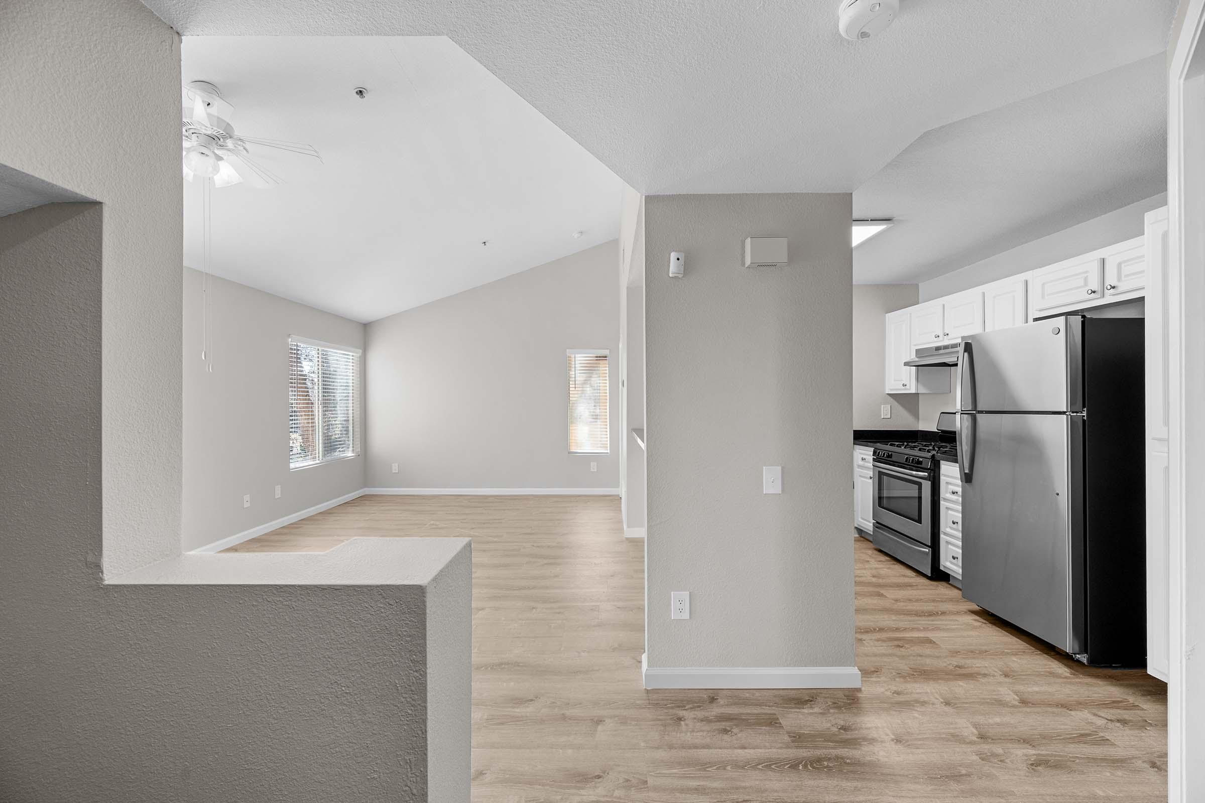 Interior view of an apartment featuring an open layout. The space includes a living area with large windows allowing natural light, a kitchen with stainless steel appliances, and light-colored walls and flooring. The vaulted ceiling creates an airy atmosphere, making the area feel spacious and inviting.