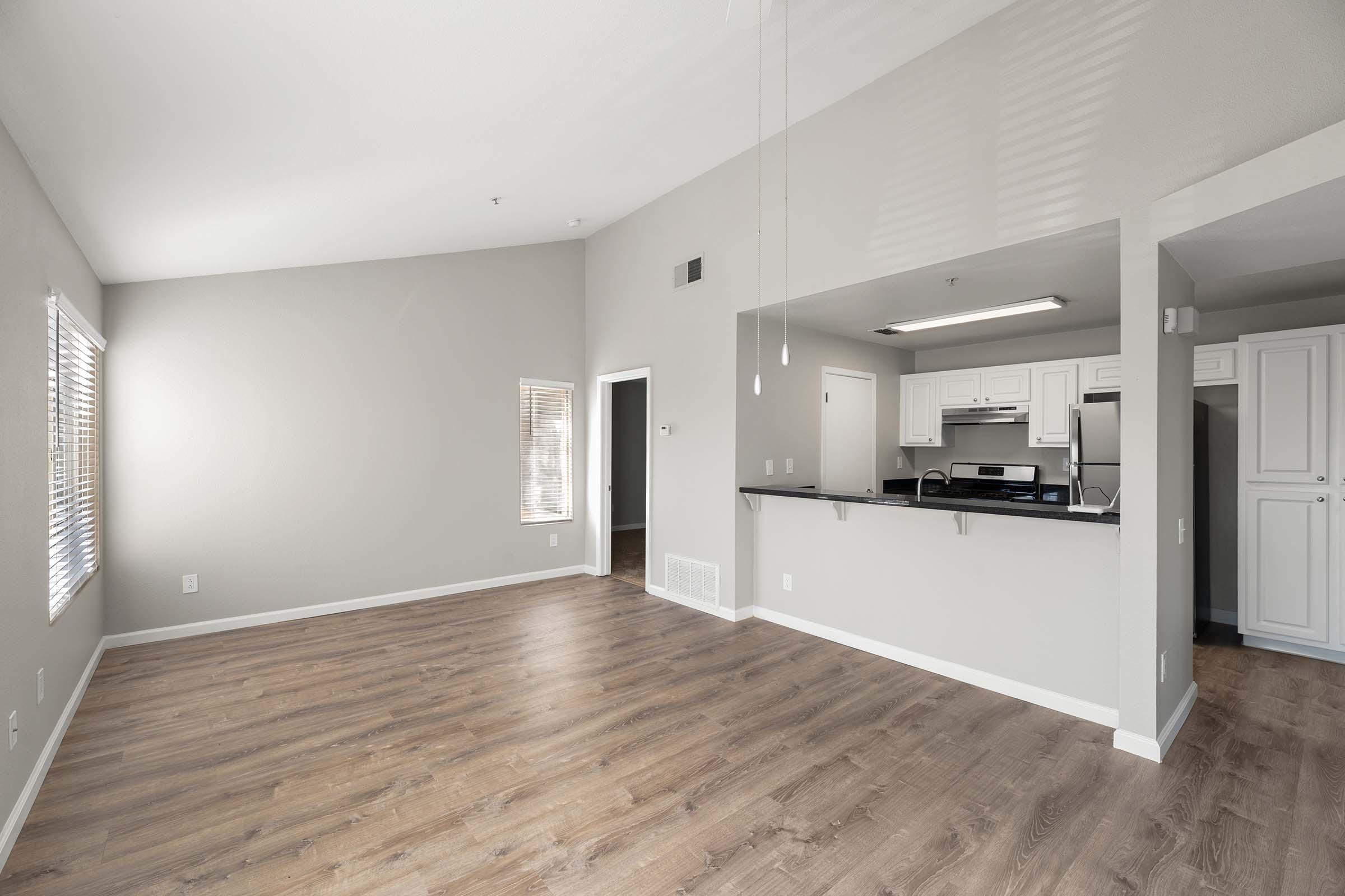 Spacious and modern living area featuring light-colored walls, large windows for natural light, and sleek wood flooring. The open layout includes an adjacent kitchen with white cabinets, stainless steel appliances, and a breakfast bar, creating a warm and inviting atmosphere.