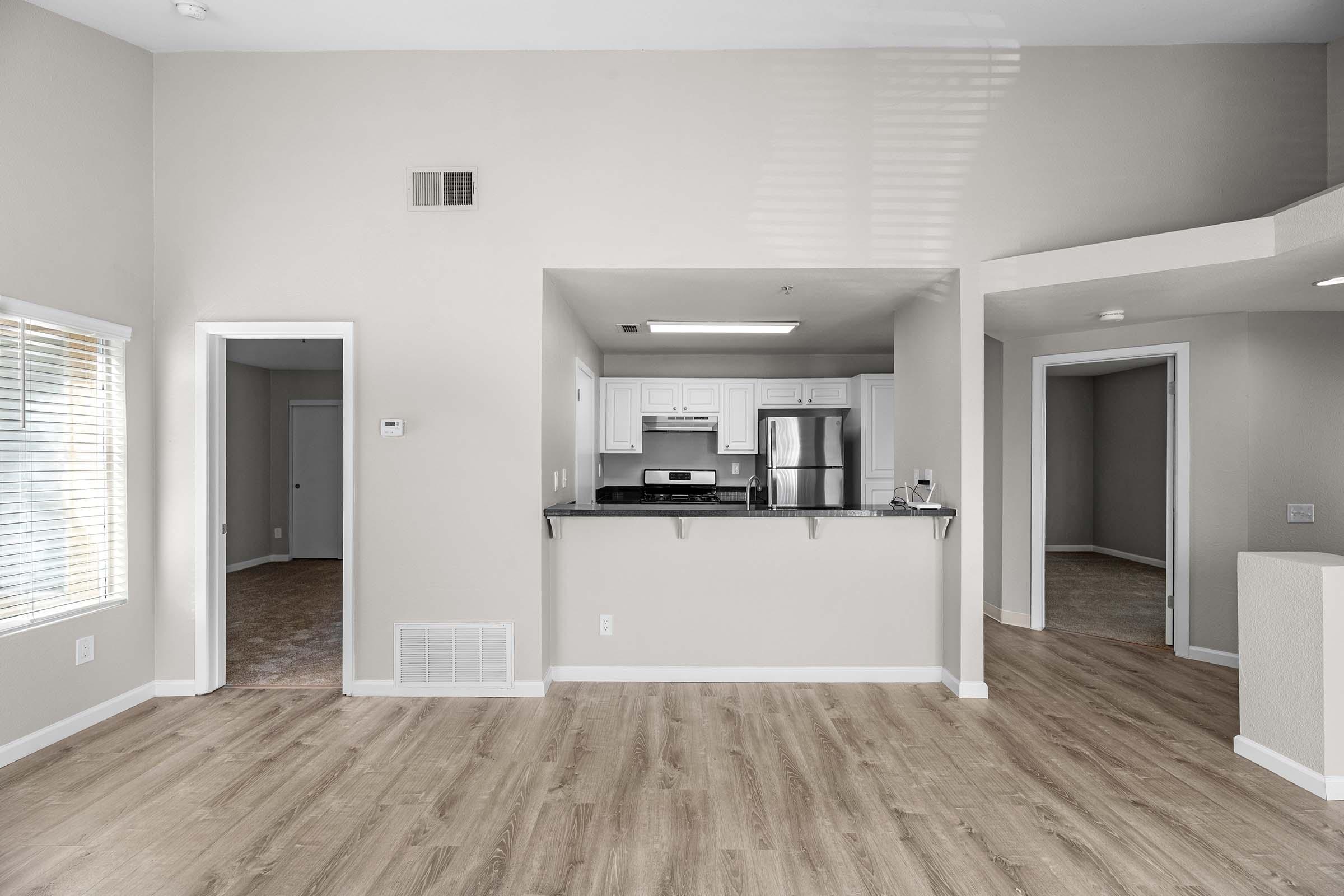 Interior view of a modern, open-concept living space featuring light-colored walls, laminate flooring, and an adjoining kitchen. The kitchen has white cabinetry, stainless steel appliances, and a breakfast bar. Natural light enters through windows, creating a bright ambiance. There are multiple doorways leading to other rooms.