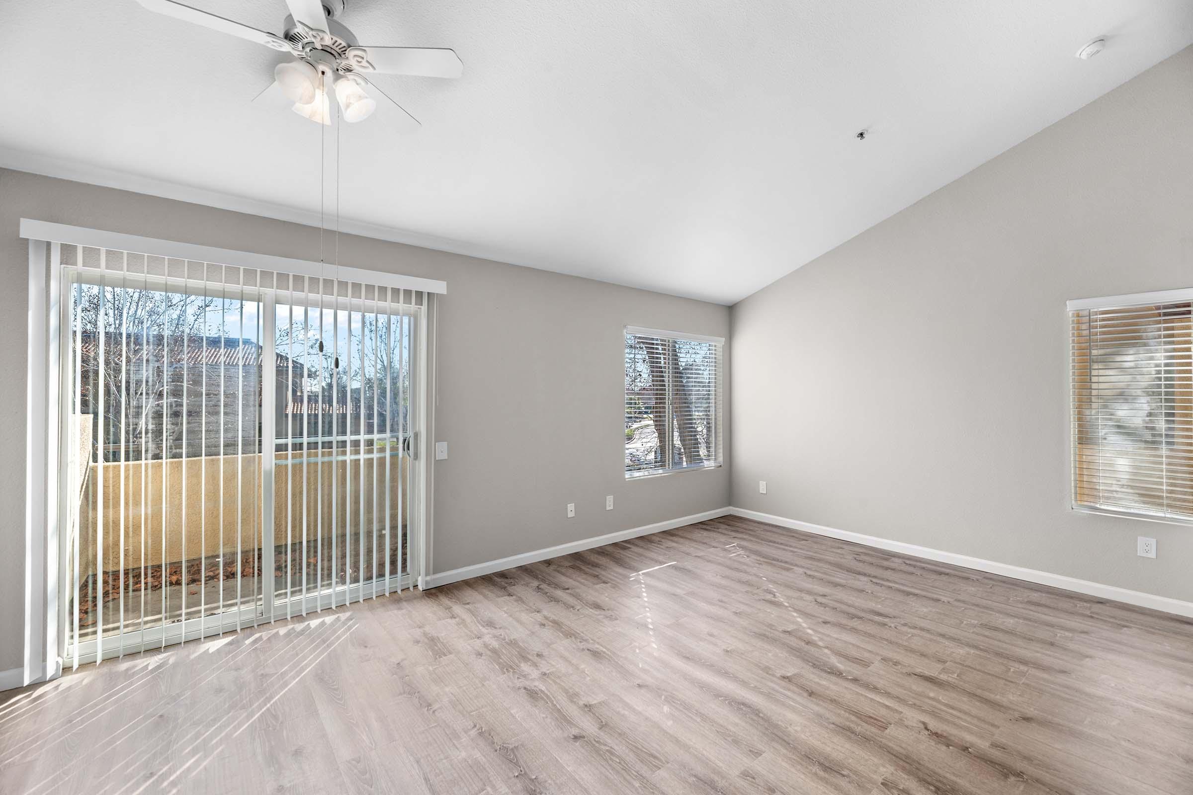 Spacious living room with high ceilings, featuring large windows with blinds, a ceiling fan, and natural light. A sliding glass door leads to a balcony, and the floor is finished with light-colored laminate wood. The walls are painted in a neutral tone, creating a warm and inviting atmosphere.