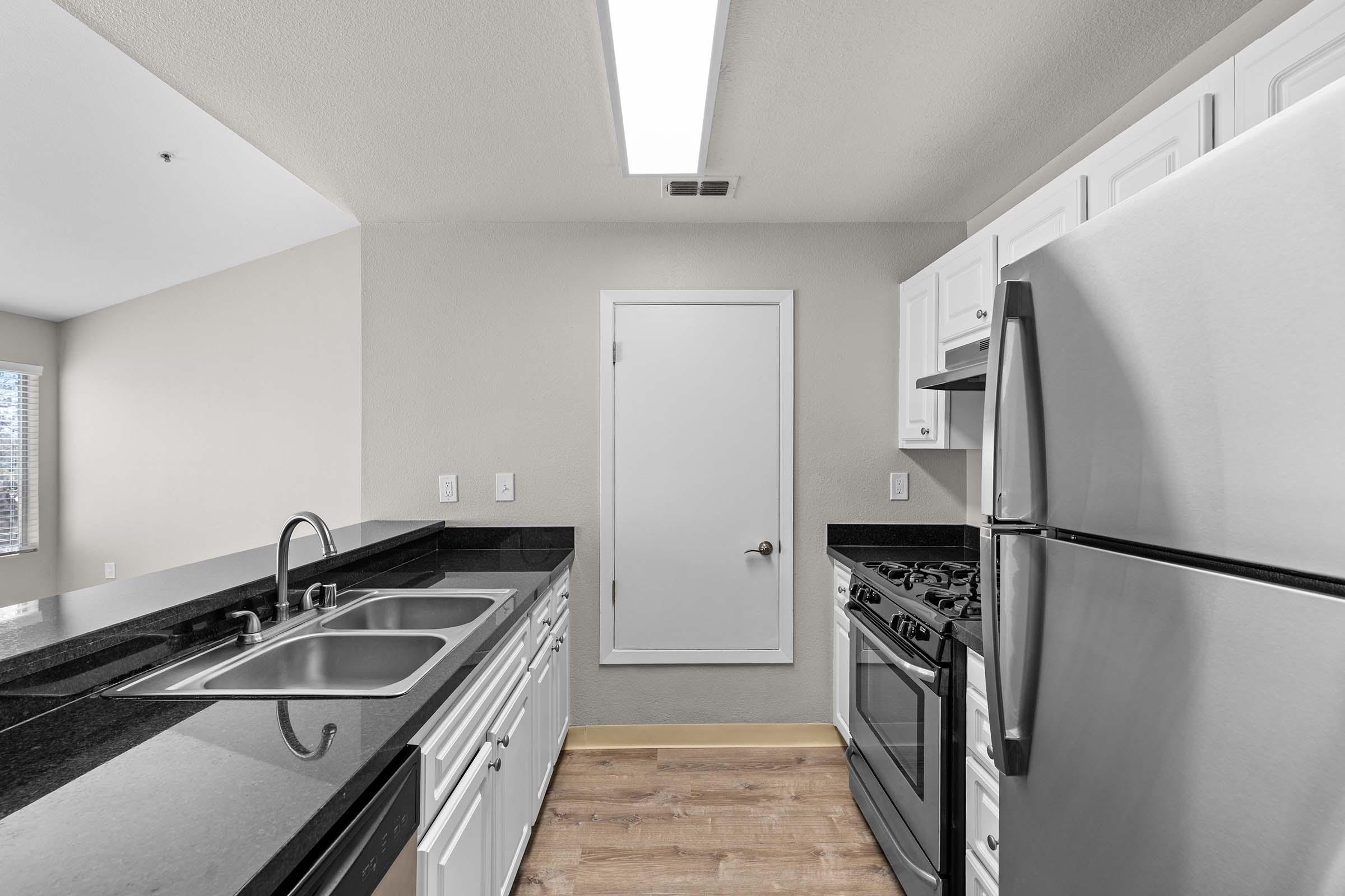 Modern kitchen with stainless steel appliances, including a refrigerator and stove. It features white cabinets, black countertops, and a double sink. Natural light comes in through a window, and the flooring is wood-like. A door leads to another room, enhancing the spacious feel.