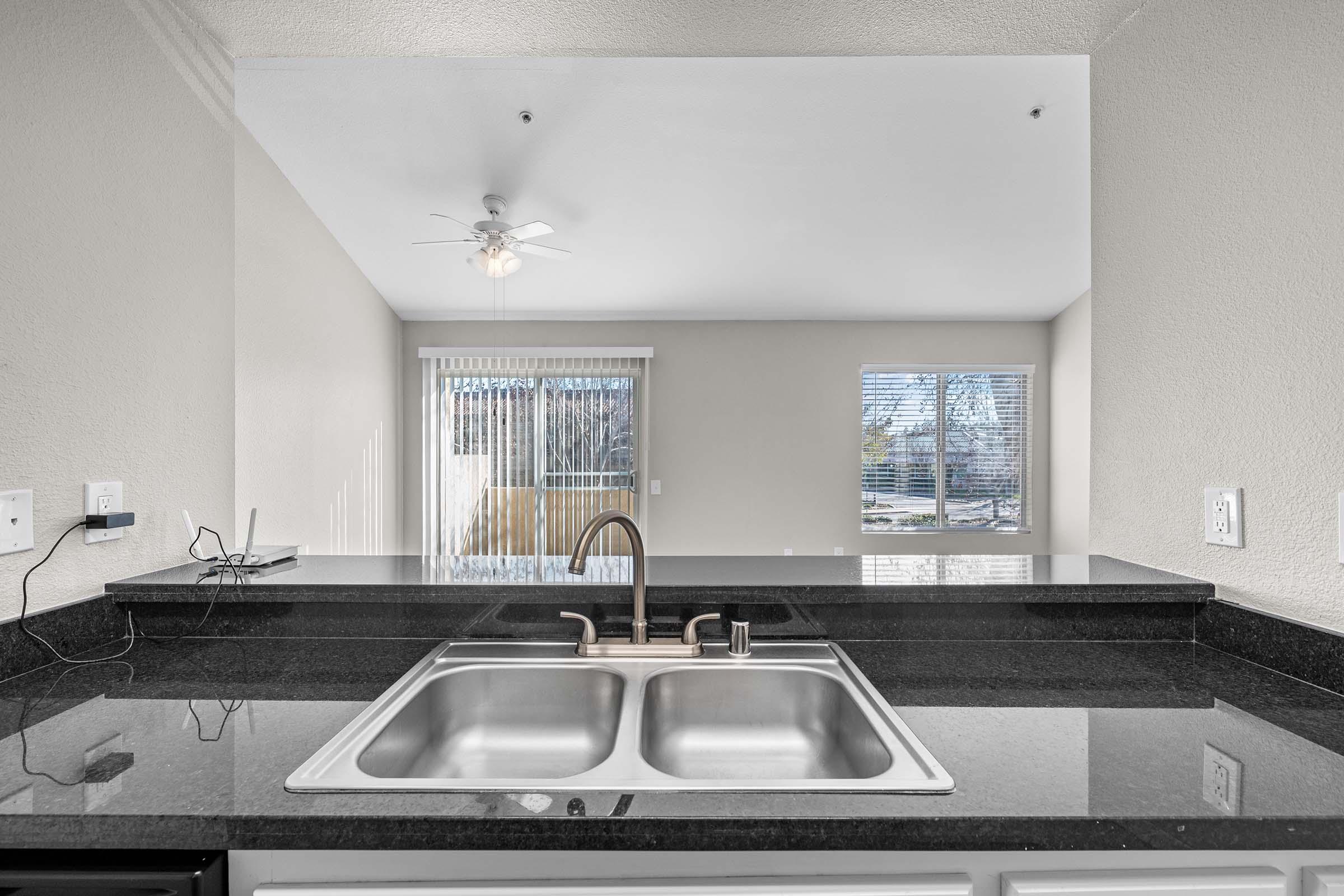 A kitchen countertop with a double sink, featuring a modern faucet. The background shows a living area with large windows, allowing natural light to enter, and a ceiling fan above. The walls are painted in a light color, creating a spacious atmosphere.