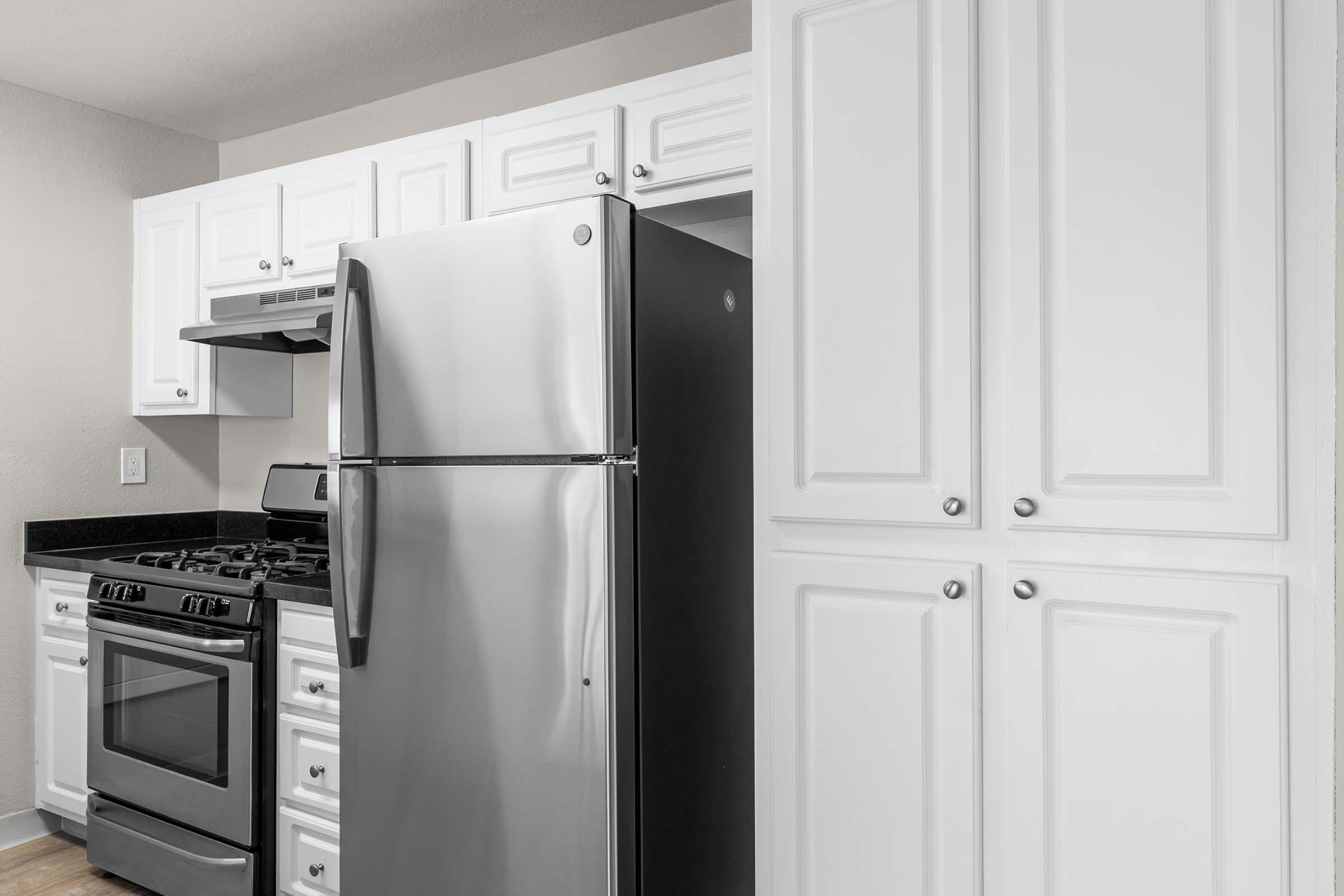 Modern kitchen featuring white cabinetry, a stainless steel refrigerator, and a black oven and stovetop. The countertop is dark, contrasting with the white cabinets. The kitchen is well-lit with a neutral wall color, providing a clean and contemporary look.