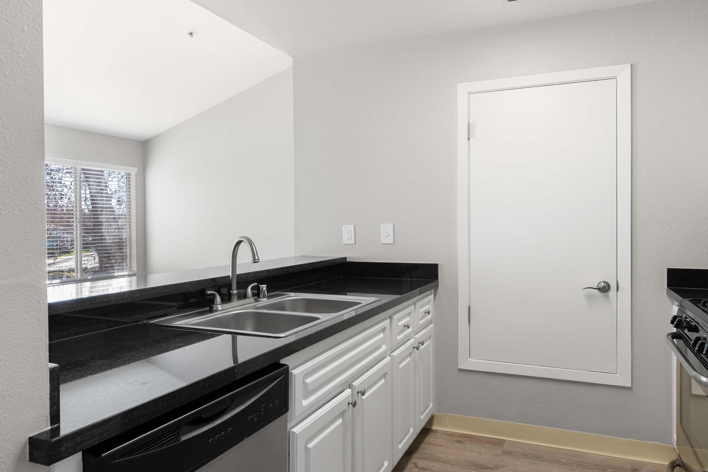A modern kitchen featuring a dark granite countertop, stainless steel sink, and white cabinetry. To the right, a door leads to another room, and a window lets in natural light. The flooring is light-colored, adding to the contemporary aesthetic.