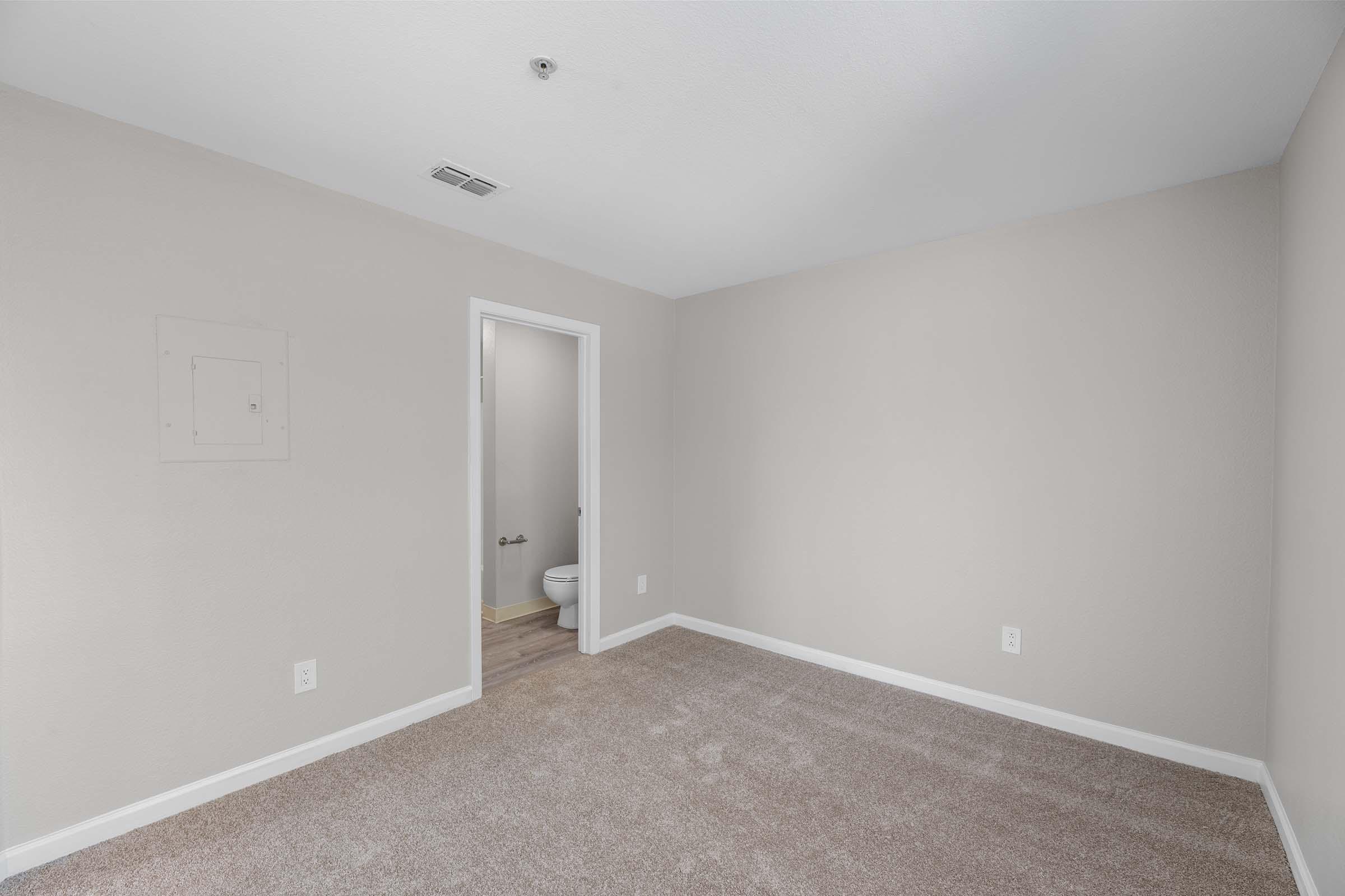 Empty room with light beige walls and neutral carpet. A doorway leads to a bathroom in the background. There is an electrical panel on one wall, and the room appears to be well-lit with natural light.