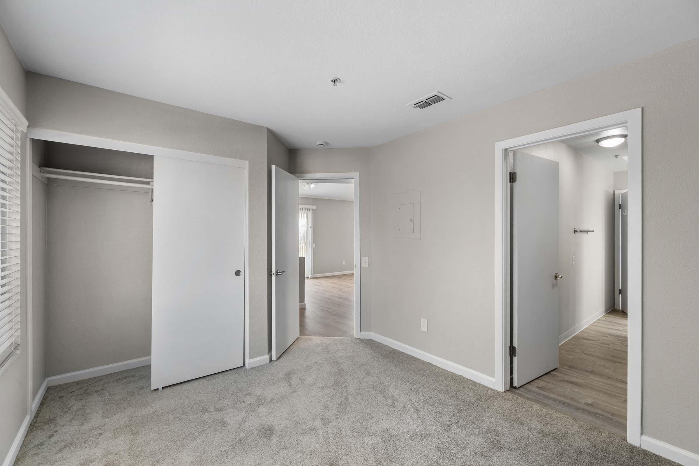 A view of a modern, empty bedroom featuring light gray walls and carpeting. There are two doors leading to other rooms and a closet with sliding doors. Natural light filters in through a window with blinds, illuminating the space. The room is well-maintained and spacious.