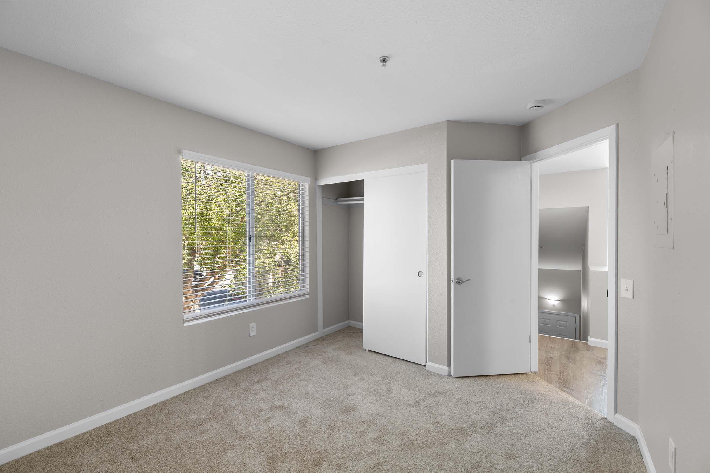 A well-lit, empty bedroom featuring light beige walls and carpeted flooring. It has a window with blinds allowing natural light, a closet, and a partially open door leading to another area. The overall appearance is clean and modern, suitable for furnishing.