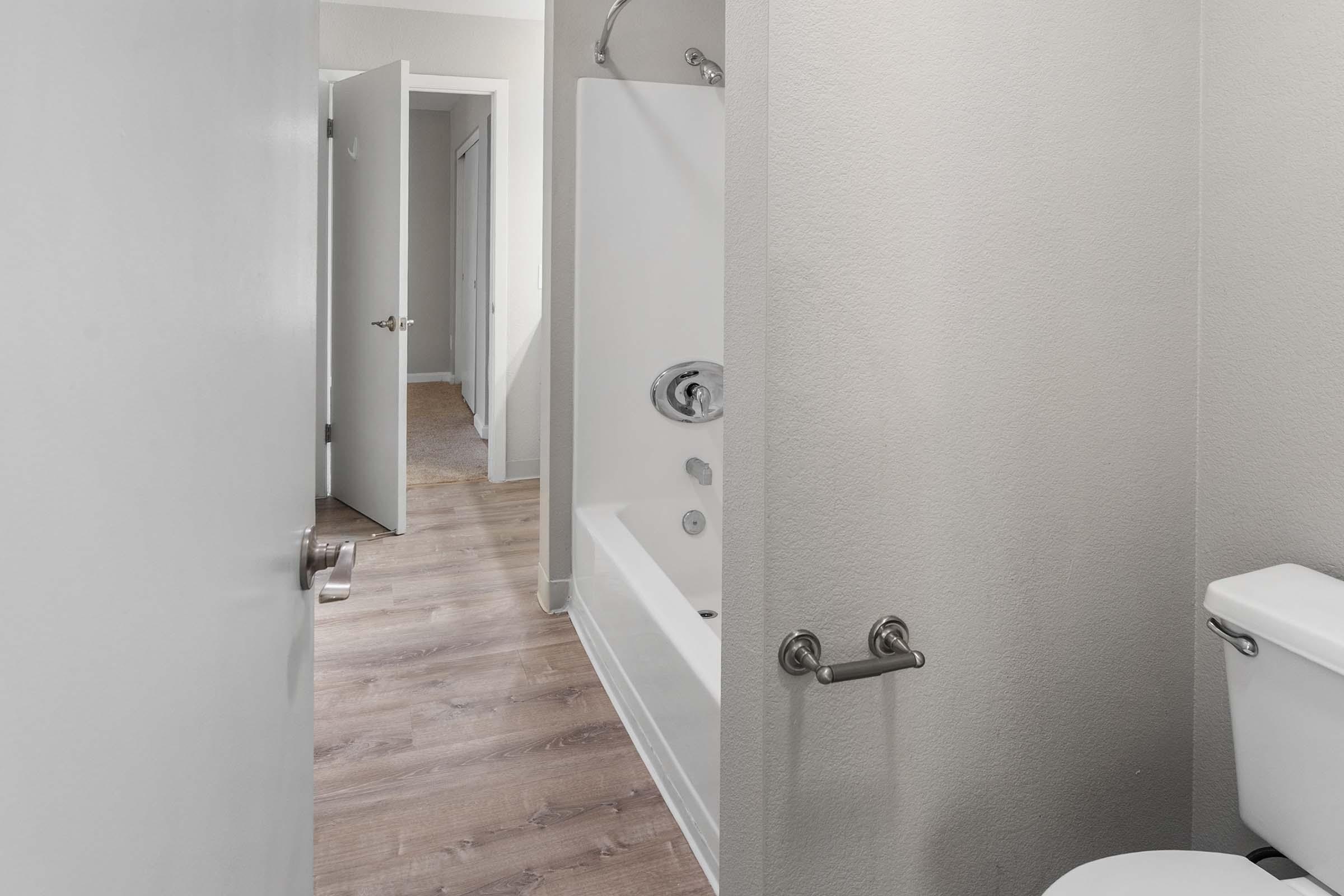A clean, modern bathroom featuring a bathtub with a shower fixture, a toilet, and neutral-colored walls. An open door leads to a hallway with a visible second door and wooden floor, enhancing the spacious feel of the area. The overall design is minimalist and contemporary.