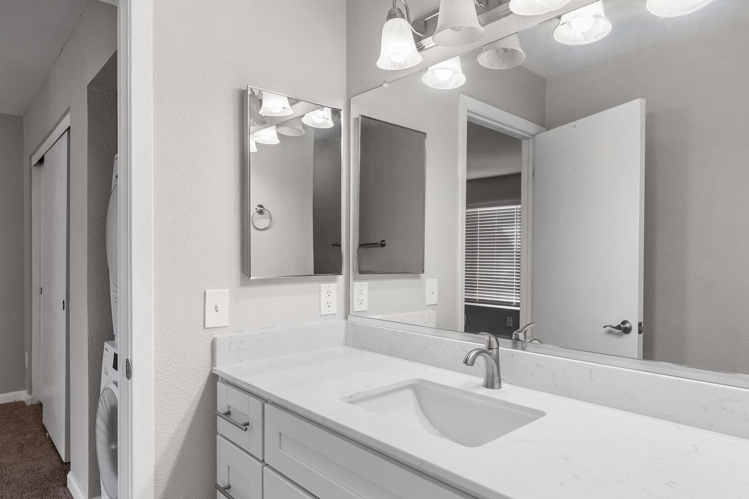 A modern bathroom featuring a white marble countertop with a sink, a large mirror, and overhead lighting. The walls are painted in neutral tones, and there is a door leading to another room along with a glimpse of a washing machine on the left. Natural light filters in through a window.