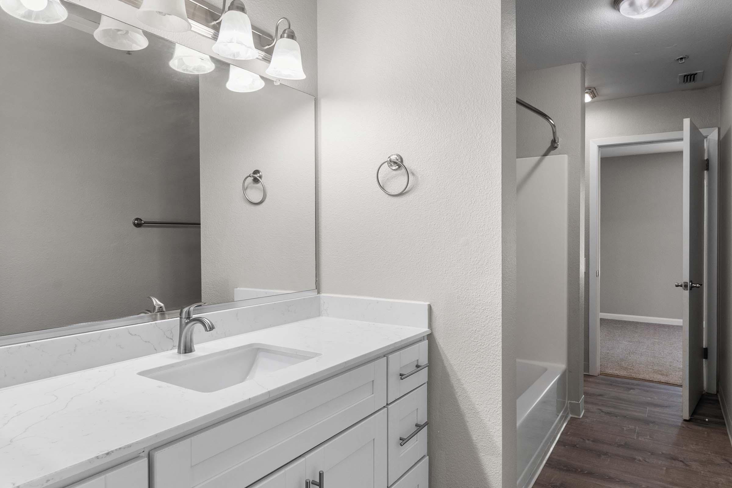 A modern bathroom featuring a white vanity with a sink, stylish light fixtures above a mirror, and a separate shower area. The walls are light-colored, and the flooring is dark wood. A doorway on the right leads to another room, and there's a towel ring on the left wall.