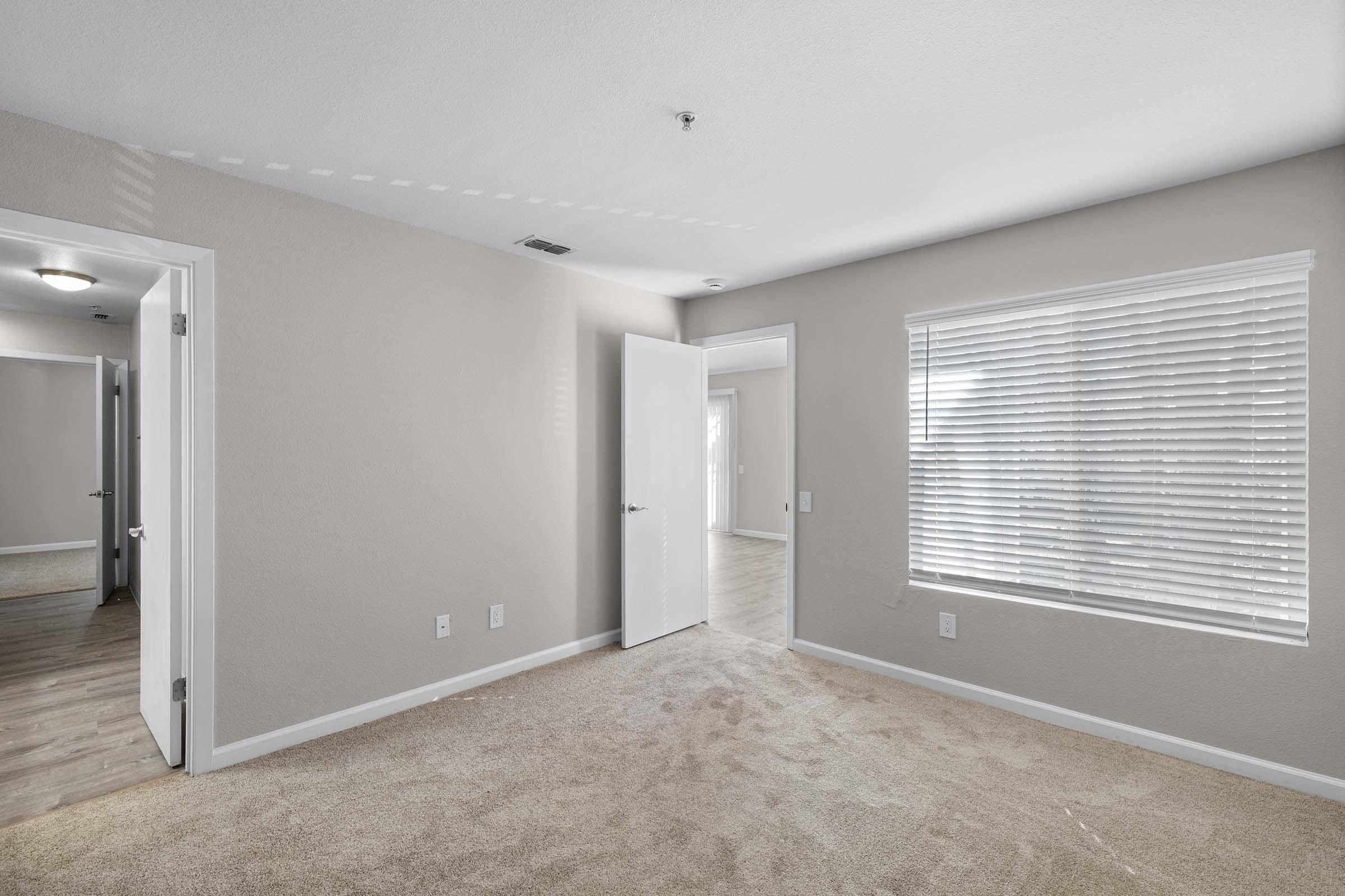 Empty room with light beige walls and carpet. There's a window with blinds allowing natural light, and an open door leading to another room. The space appears clean and modern, suitable for furniture arrangement. Additional doorways are visible, indicating access to other areas.