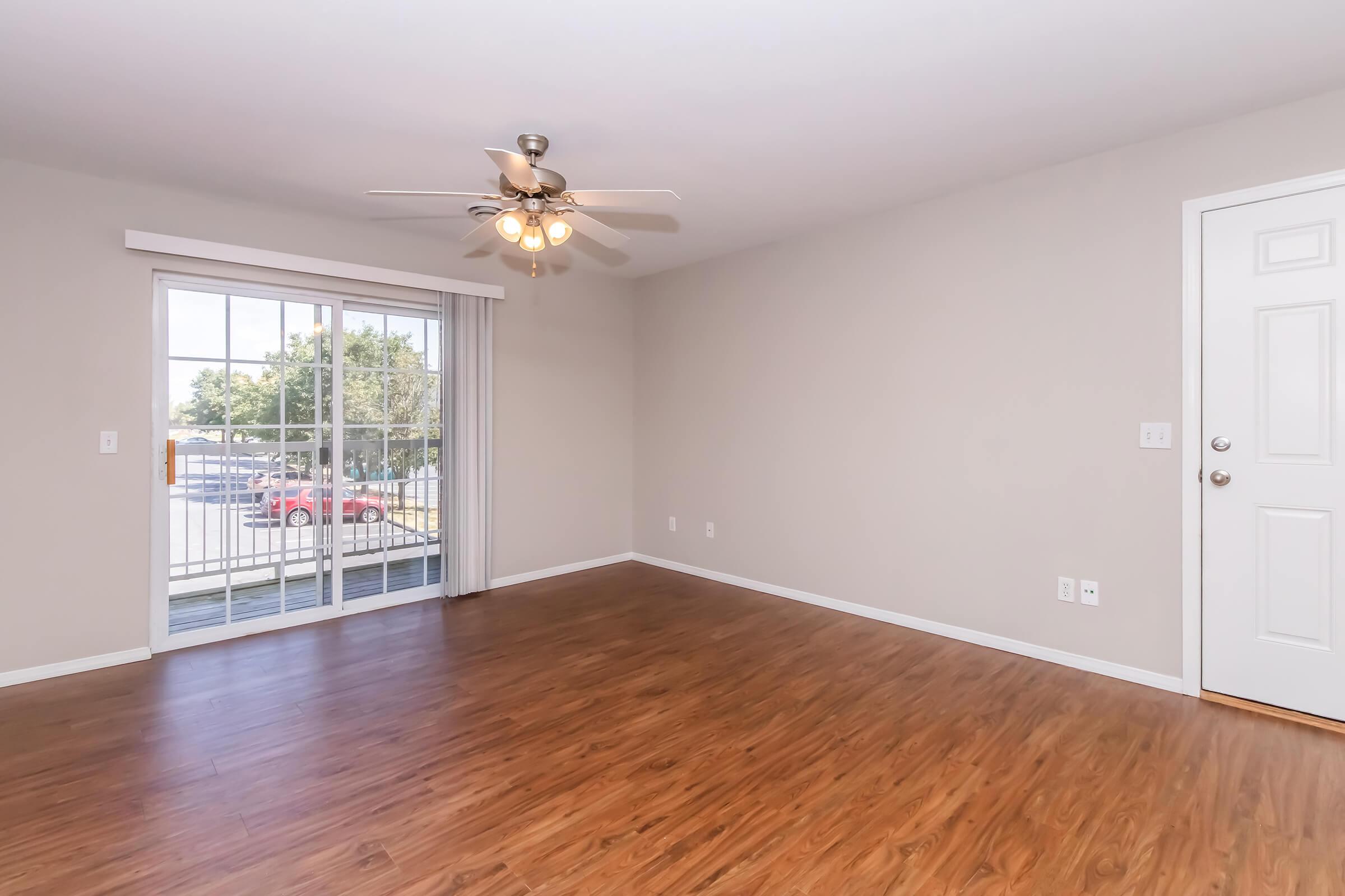 A spacious empty room with light brown laminate flooring, a ceiling fan, and a large window with white blinds. The walls are painted a light beige color. A white door is visible on the right side, leading outside. The view outside includes a glimpse of cars parked near the building.