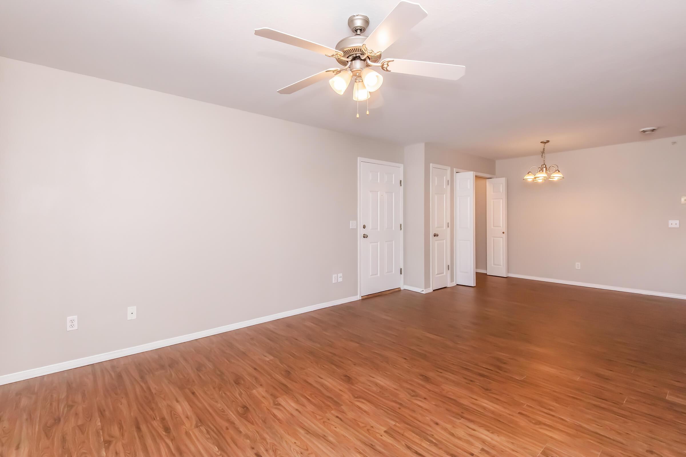 Spacious, empty living area featuring a ceiling fan with light fixtures, neutral wall colors, laminate flooring, and doorways leading to adjacent rooms. The layout is bright and inviting, ideal for personal customization.