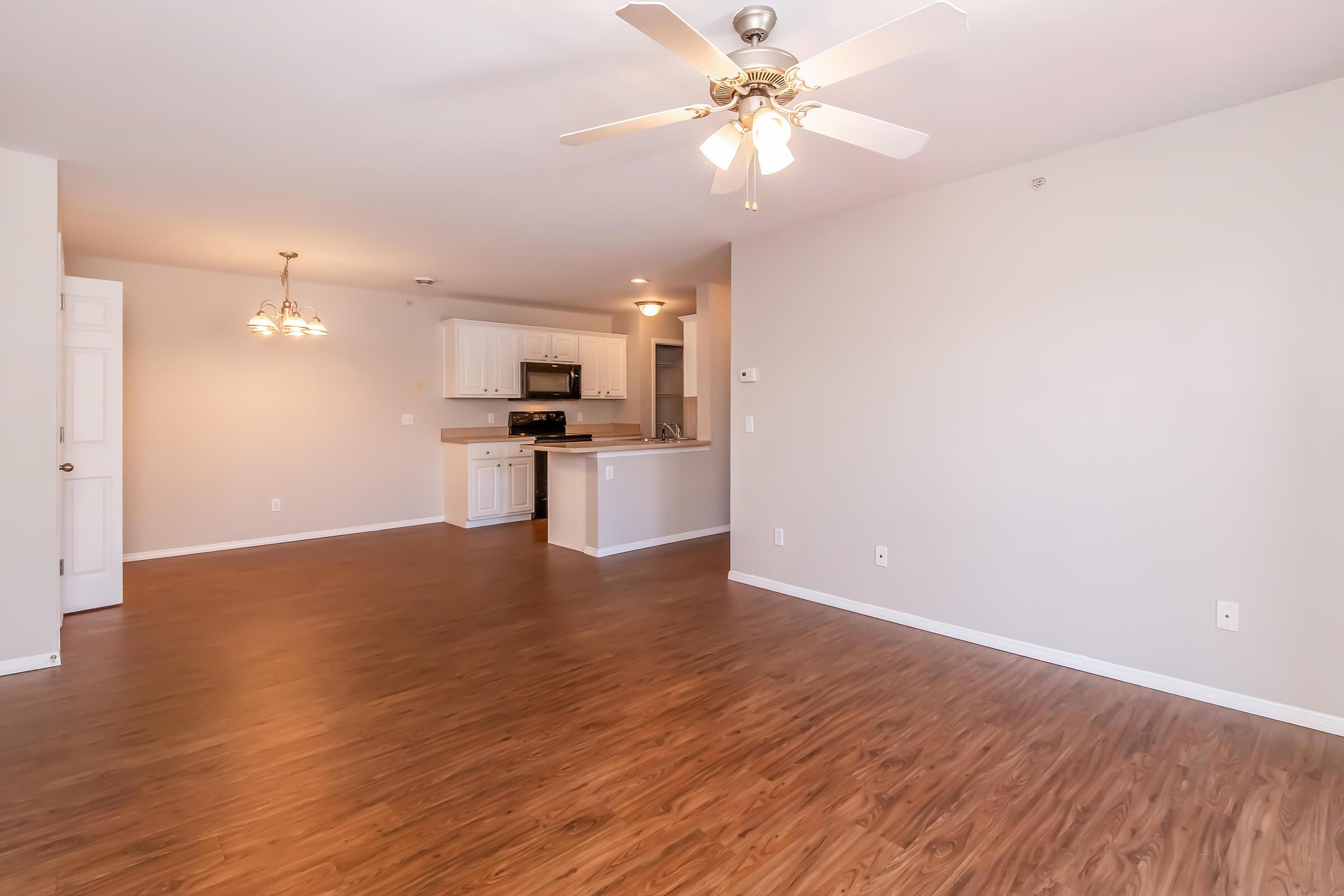 Bright and spacious living area with light-colored walls and hardwood flooring. A ceiling fan provides ventilation. The open layout leads to a modern kitchen featuring white cabinetry and appliances, with pendant lighting over the dining area. Ideal for creating a comfortable home environment.