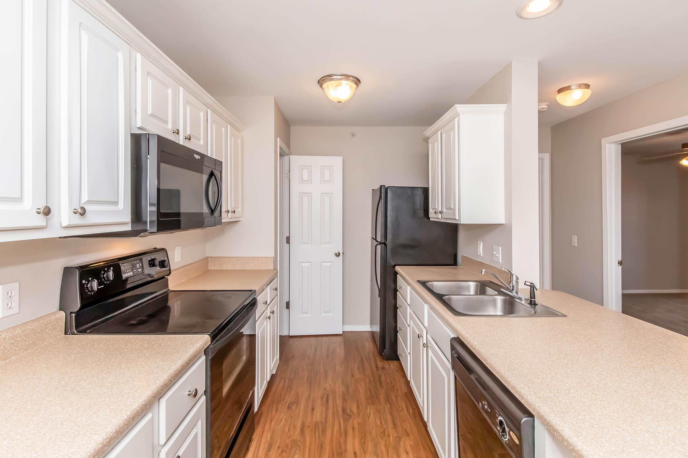 Modern kitchen featuring white cabinets, stainless steel appliances, and a light-colored countertop. Includes a black microwave, oven, and refrigerator, with a double sink. The walls are painted in a neutral shade, and there's a door leading to another room. The flooring is wood-like laminate.
