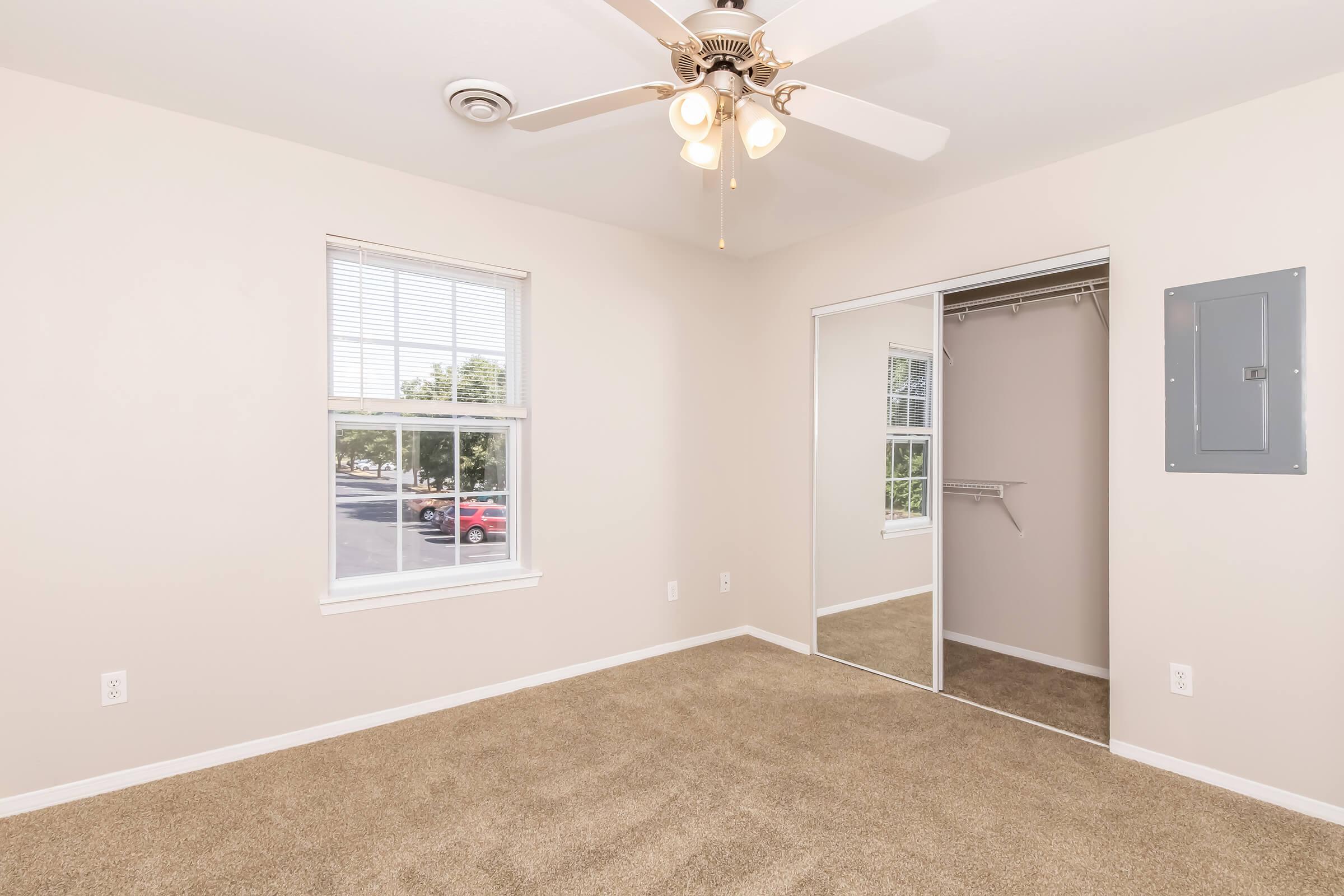Bright, empty bedroom with light beige walls and plush carpet. Features a ceiling fan with light fixtures, a window with blinds providing natural light, and a mirrored closet door. Electrical panel mounted on the wall and a spacious feel throughout the room.