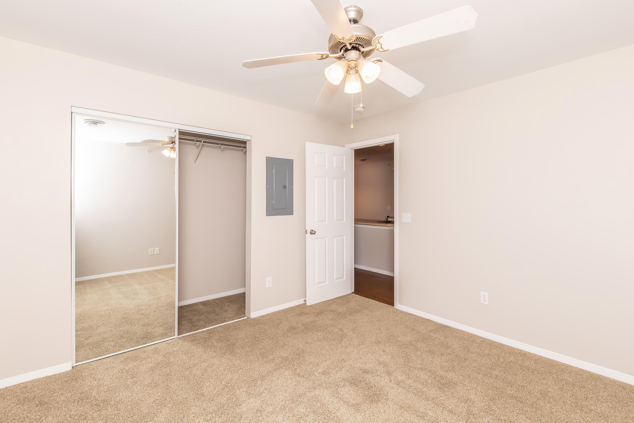 Empty bedroom with light beige walls and carpet. Features a ceiling fan with lights, a mirrored closet with sliding doors, and a closed door leading to another room or space. The room is well-lit and spacious, creating a welcoming atmosphere.