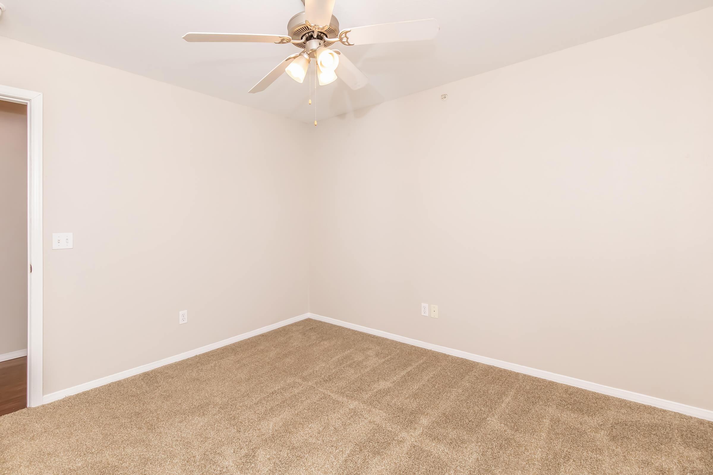 A spacious room featuring light-colored walls and a beige carpet. A ceiling fan with five blades hangs from the ceiling, providing potential cooling. The room is empty, with no furniture visible, and has a door on the left side. The overall ambiance is bright and inviting.