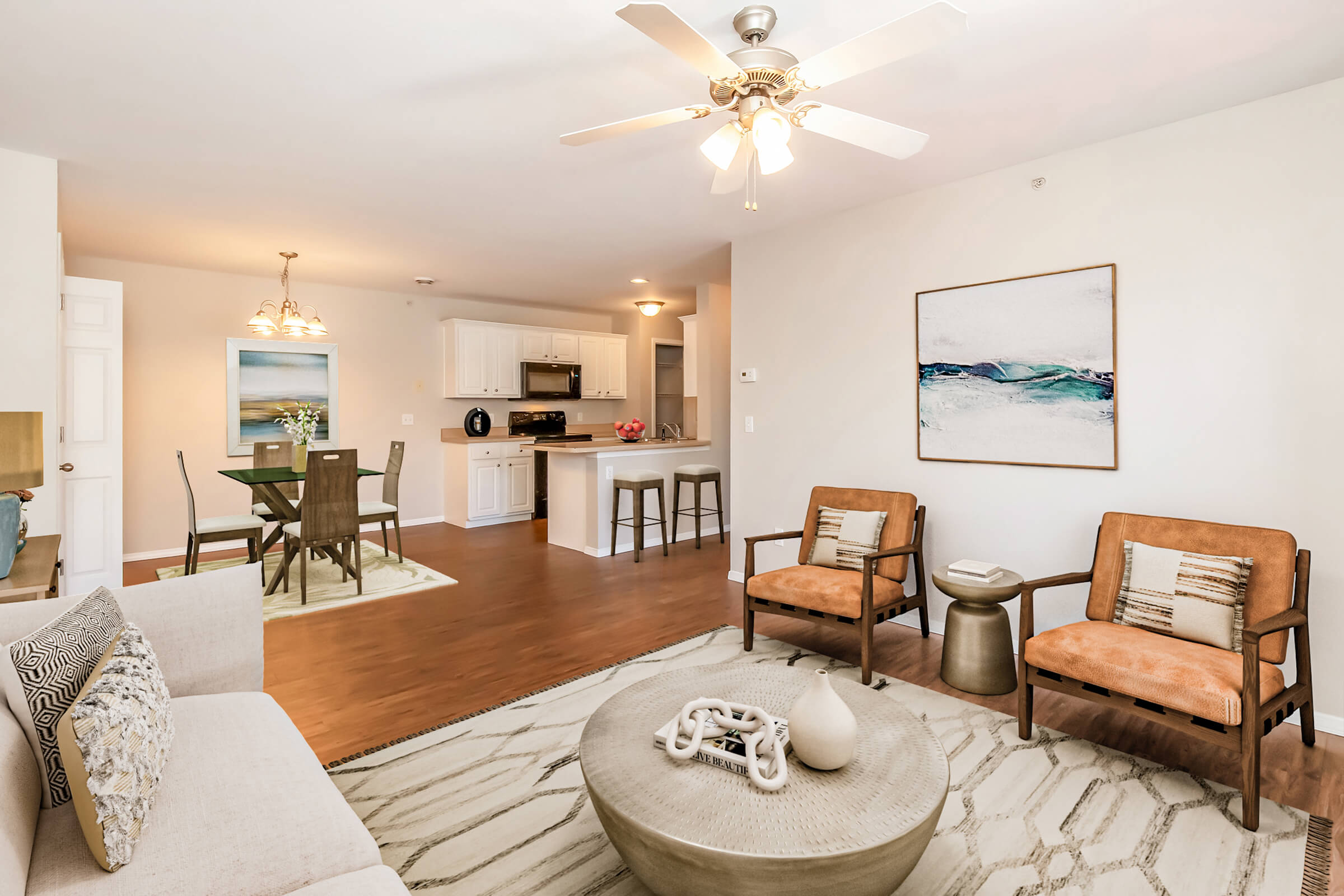 A modern living room and dining area featuring two brown armchairs, a light-colored sofa, a circular coffee table, and a decorative centerpiece. The space has a ceiling fan, light wood flooring, and an open kitchen in the background with white cabinetry. A large piece of abstract wall art adds a touch of color.