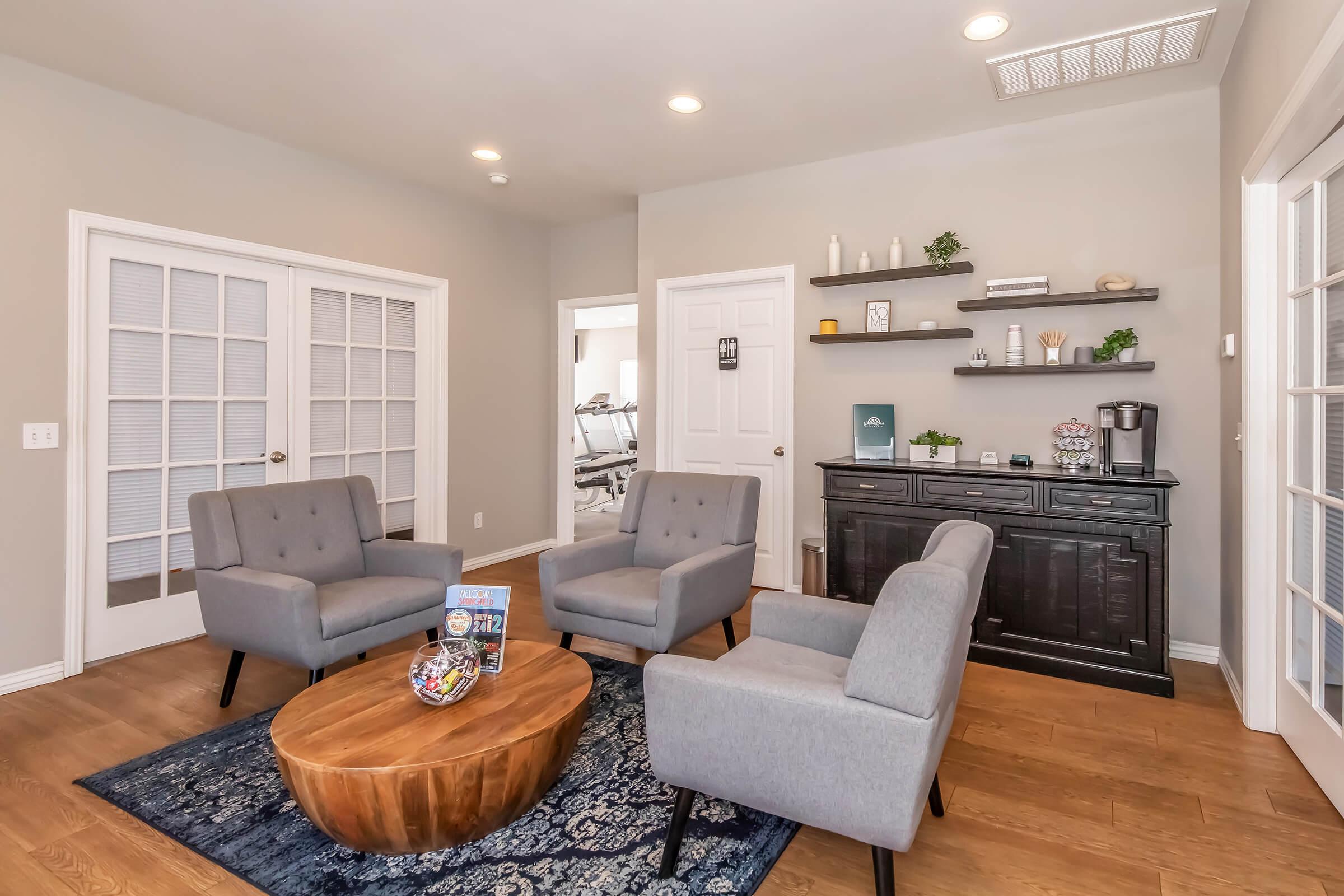 A modern interior space featuring four gray armchairs arranged around a round wooden coffee table. There is a black cabinet against the wall, decorated with plants and accessories. The walls are light gray, and double doors lead to a gym area visible in the background. Natural light brightens the room.