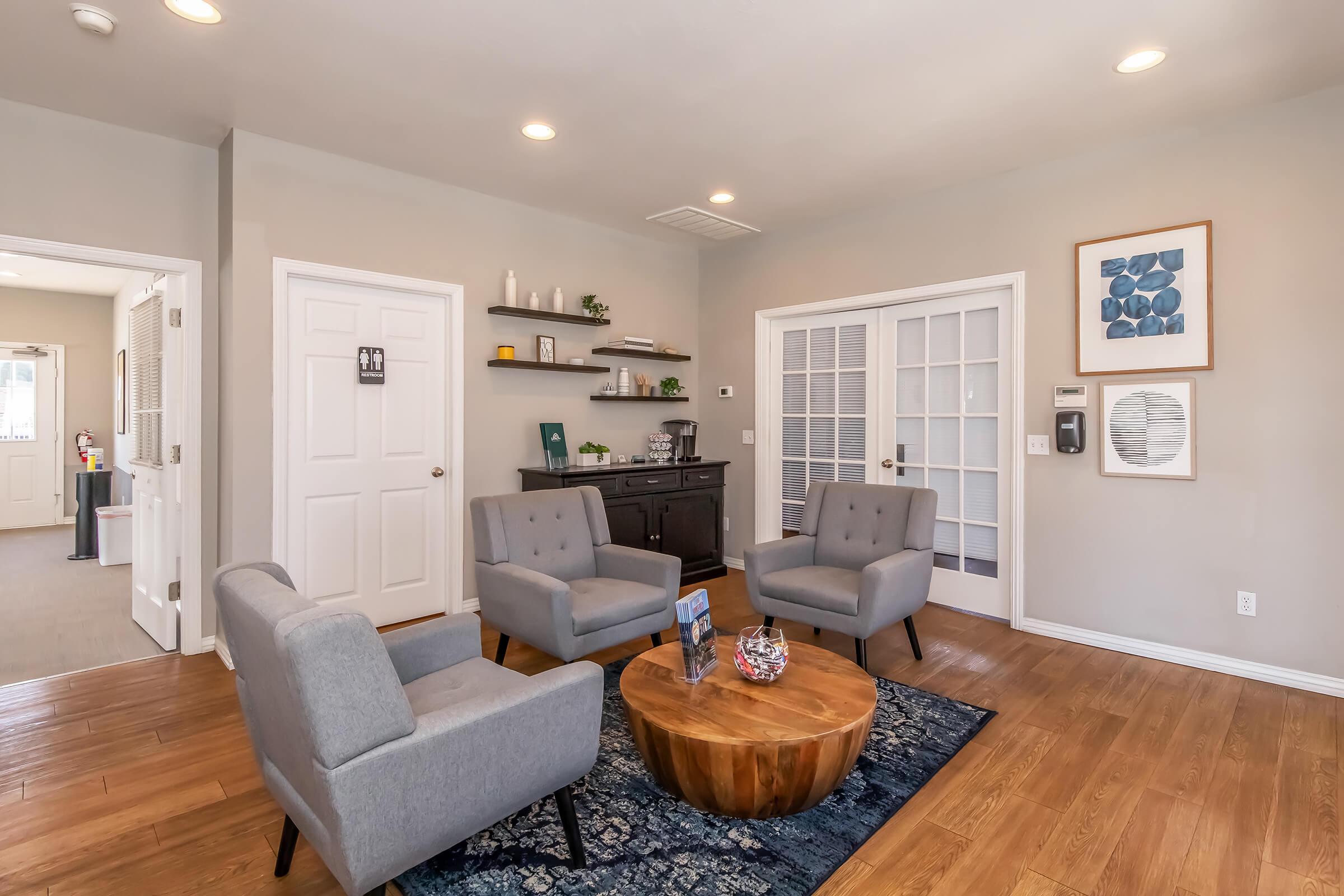 A cozy waiting area with two light gray armchairs and a round wooden coffee table on a blue rug. A dark wooden cabinet against the wall contains decorative items. There are two doors leading to other rooms, and a wall art piece featuring geometric shapes. Soft lighting creates a welcoming atmosphere.