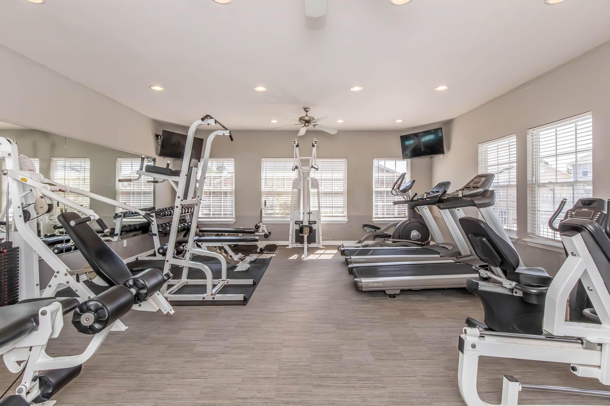 A well-lit gym featuring various exercise equipment, including weight machines, treadmills, and ellipticals. Large windows provide natural light, and there are multiple ceiling lights. The floor is wood-like, creating a warm atmosphere for workouts.