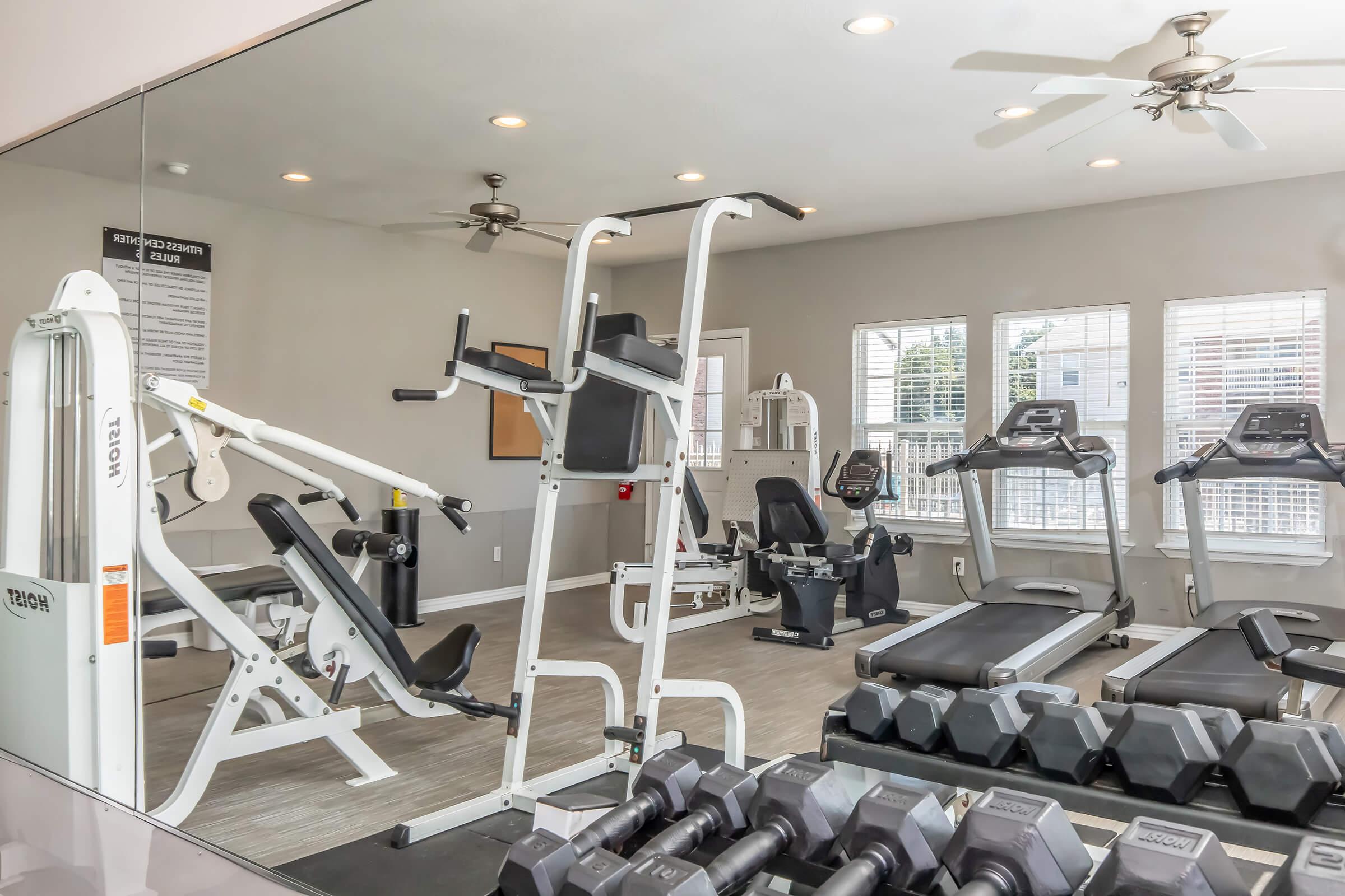 A well-equipped gym with various exercise machines including a bench press, treadmill, and stationary bikes. Free weights are neatly arranged on a rack. Bright windows let in natural light, and the room features ceiling fans for ventilation. The overall atmosphere is clean and inviting.
