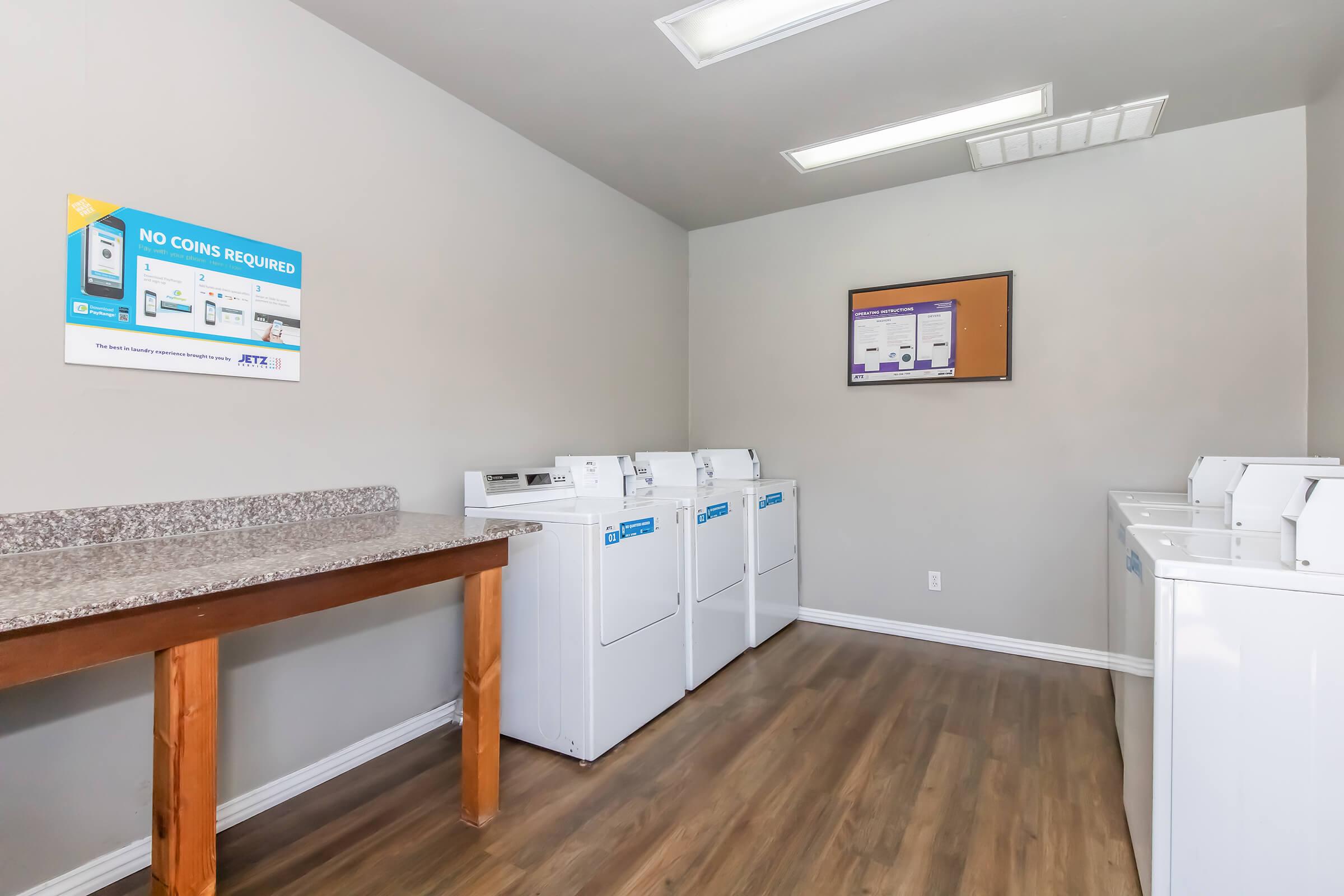 A clean laundry room featuring several white washing machines lined against a gray wall. There is a wooden table on one side, and a sign on the wall indicating that no coins are required for usage. The floor is made of light-colored wood, and the room is well-lit with overhead lights.