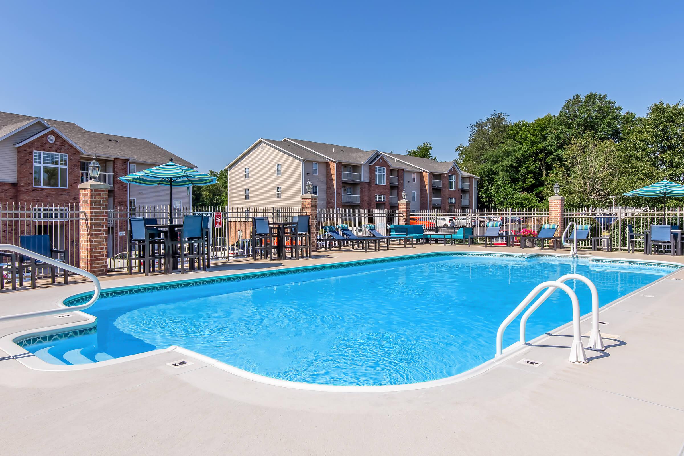 A clear blue swimming pool surrounded by concrete and lounge chairs, with tables and umbrellas nearby. In the background, there are two-story apartment buildings, and greenery is visible on the edges under a clear blue sky.