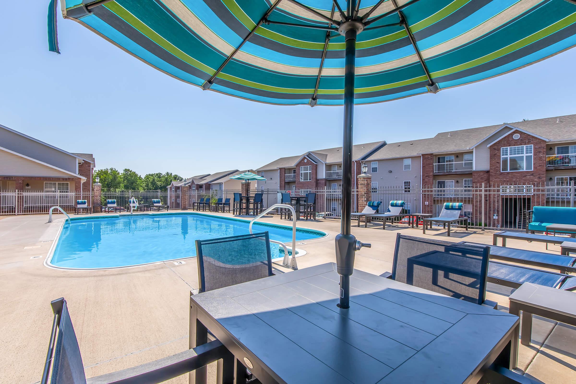 A swimming pool area with lounge chairs and patio furniture under a striped umbrella. The pool is surrounded by residential buildings, and clear blue skies overhead create a sunny atmosphere.