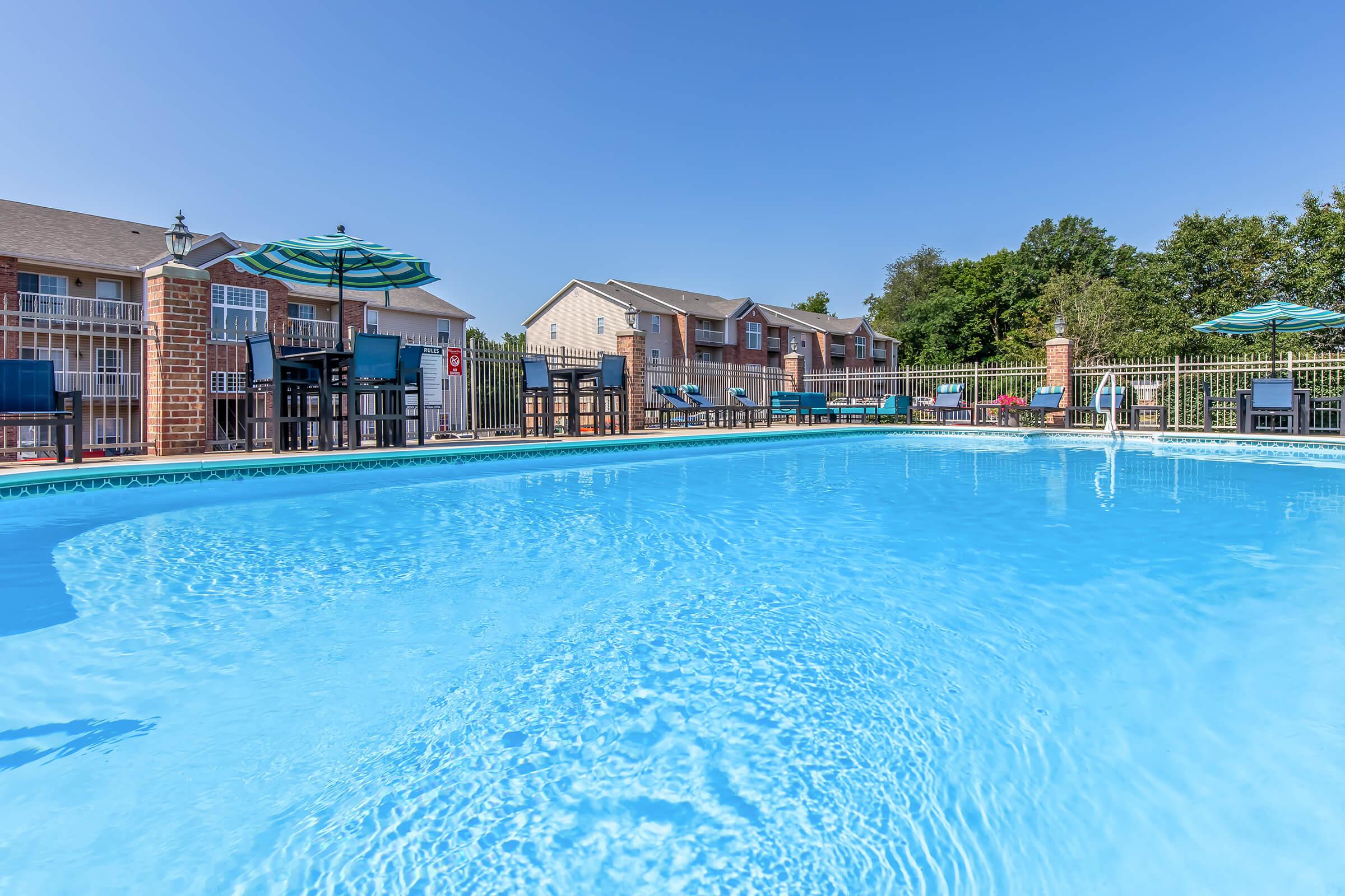 A view of a clear blue swimming pool surrounded by lounge chairs and umbrellas. In the background, there are two-story buildings with balconies and green trees, under a clear blue sky. The scene conveys a refreshing and inviting atmosphere for relaxation and leisure.