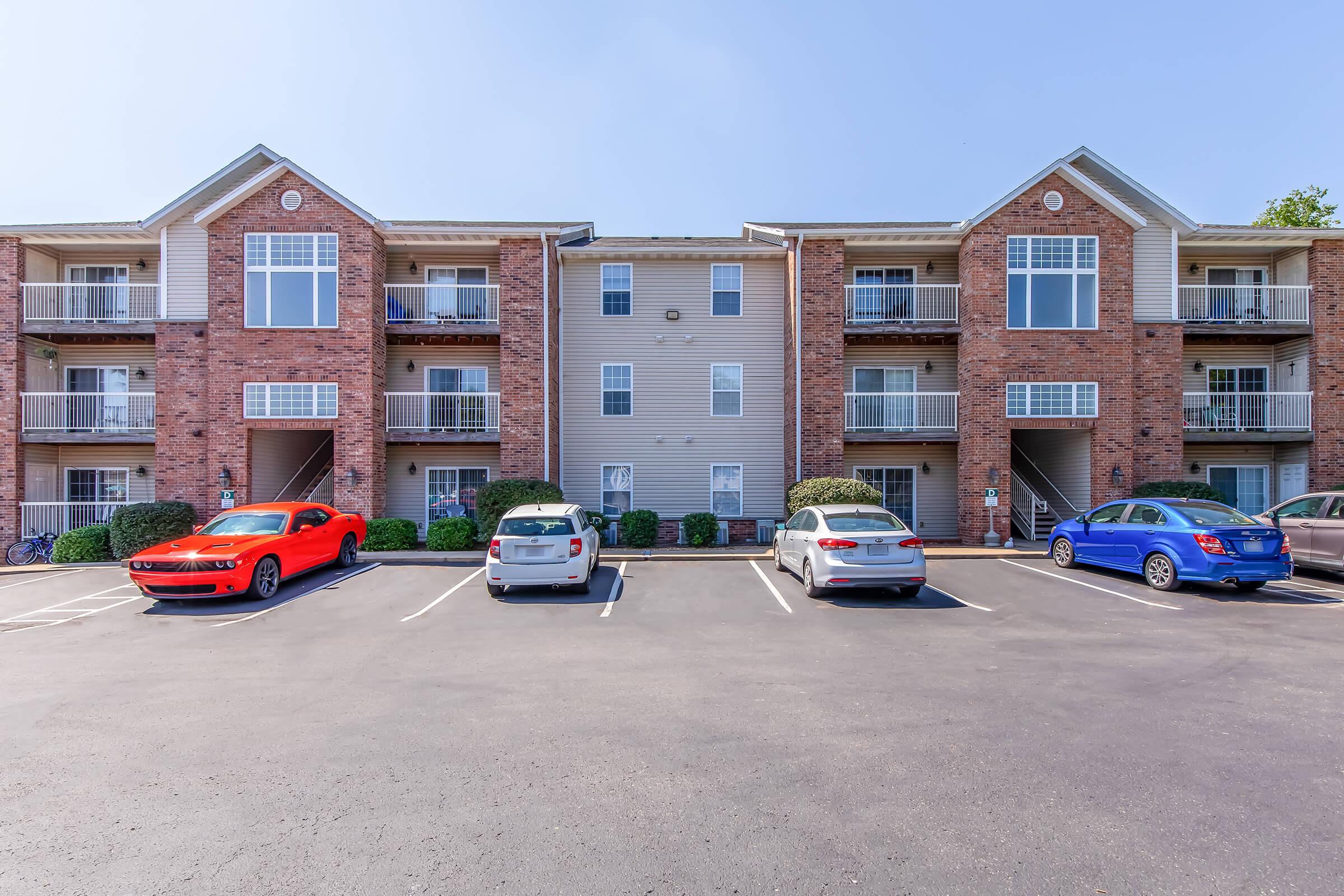 A view of an apartment complex featuring two buildings made of brick and siding. The buildings have multiple balconies and parking spaces in front. Several cars are parked in the lot, including a red sports car, a silver sedan, and a blue car. Clear blue skies are visible above.