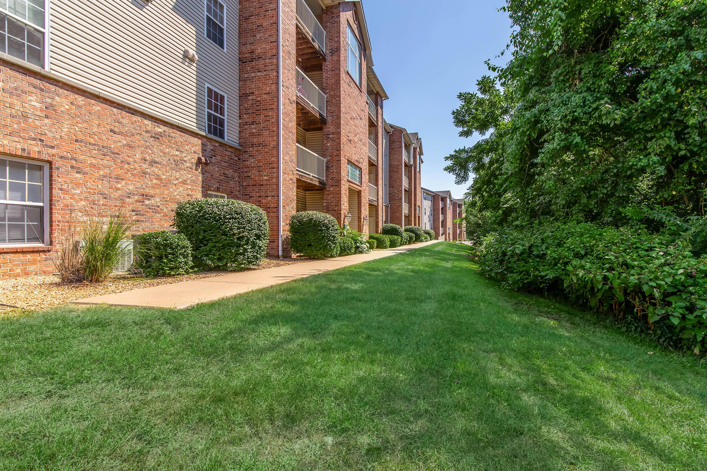 View of a landscaped pathway alongside a brick apartment building. The scene features well-maintained lawns, shrubs, and trees, creating a peaceful residential environment under a clear blue sky. The walkway leads toward additional apartment units in the background.