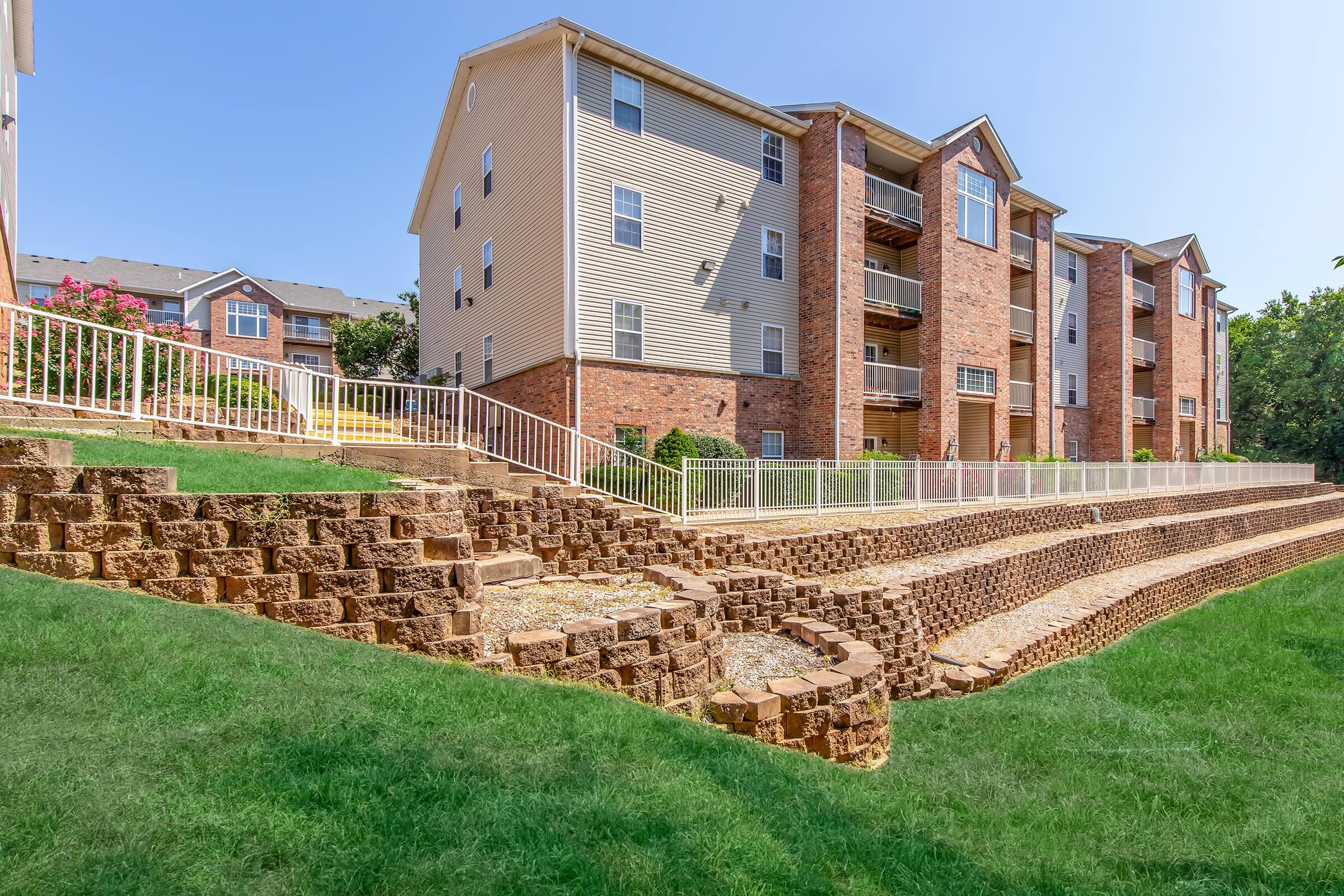 A view of a multi-story apartment building surrounded by well-maintained landscaping. In the foreground, there are tiered stone walls leading up to the building, with green grass and a clear blue sky overhead. The setting is bright and inviting, showcasing a peaceful residential environment.