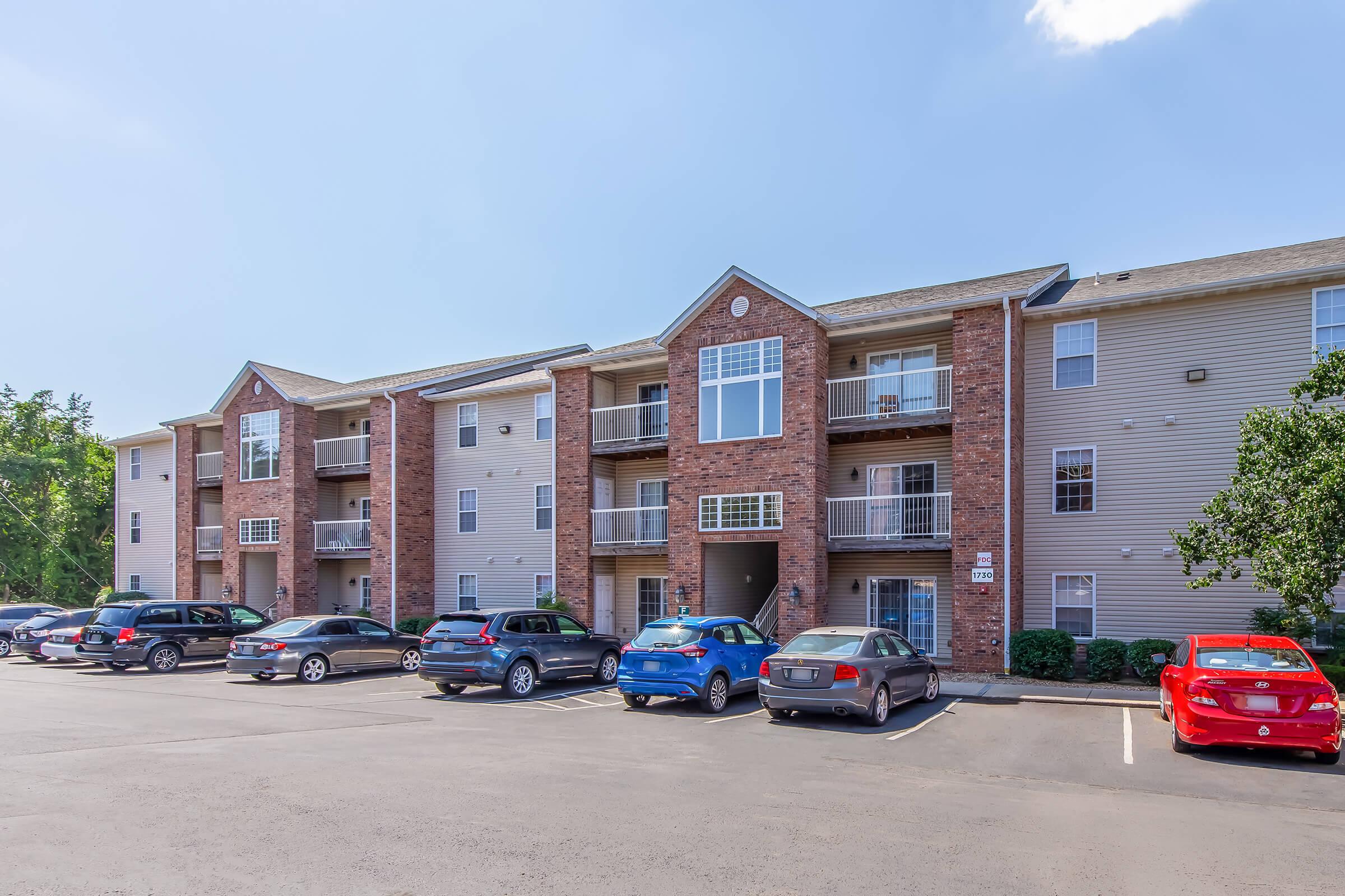 A multi-story residential apartment building with a brick façade and light-colored siding. The building features multiple balconies, a parking lot filled with various cars, and greenery in the surrounding area. The sky is clear with a few clouds.