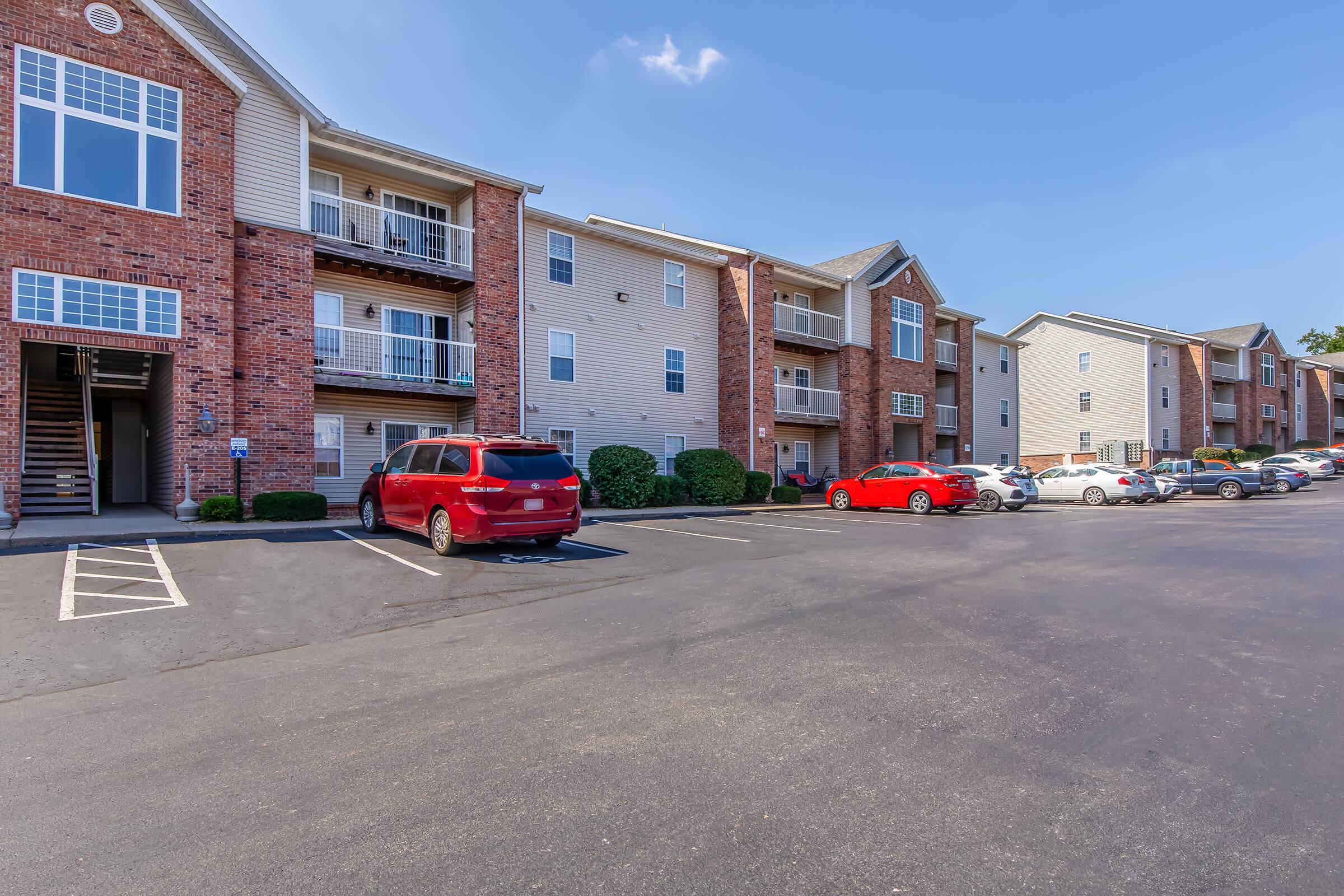 View of an apartment complex with brick and siding buildings. Several parked cars, including a red vehicle, are visible in the foreground. The sky is clear, and the overall scene suggests a well-maintained residential area.