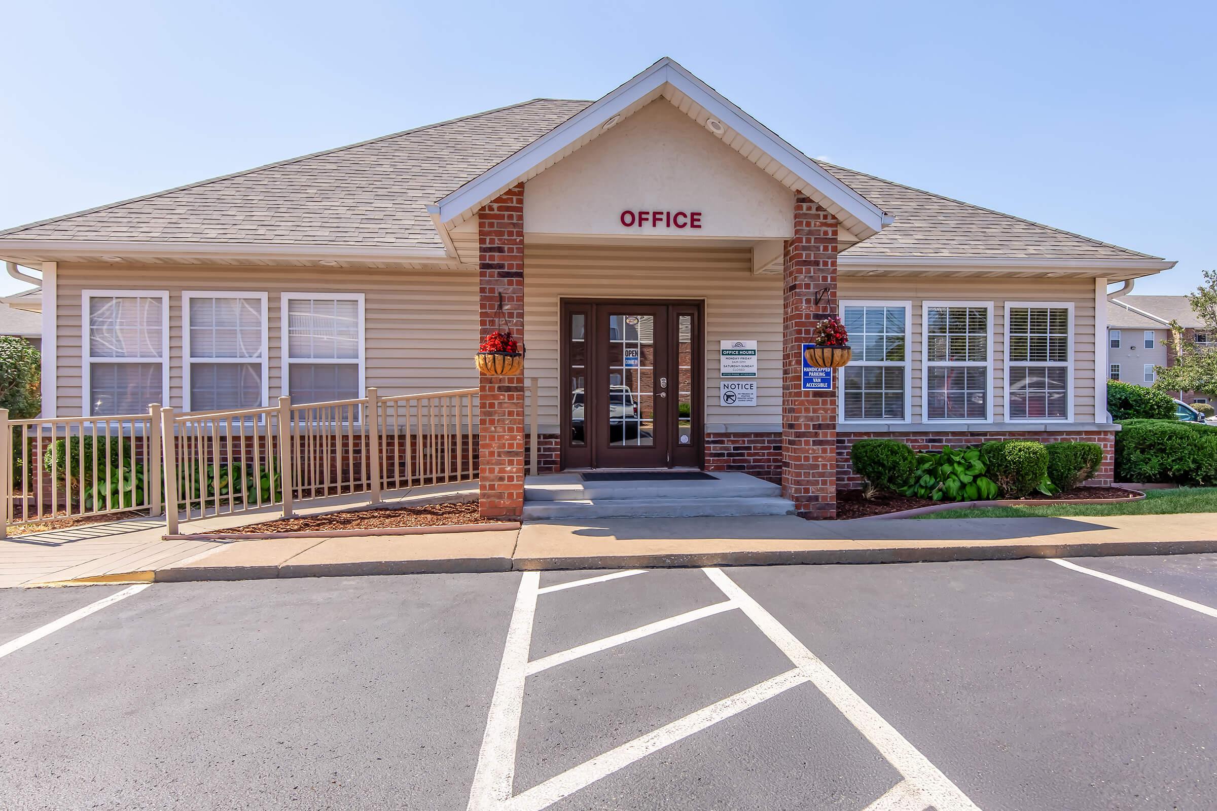 A single-story office building with a peaked roof, featuring large windows and a front porch. The word "OFFICE" is displayed prominently above the entrance. Decorative flower pots are placed on either side of the door, and there is a nearby parking area with marked spaces. The sky is clear and blue.
