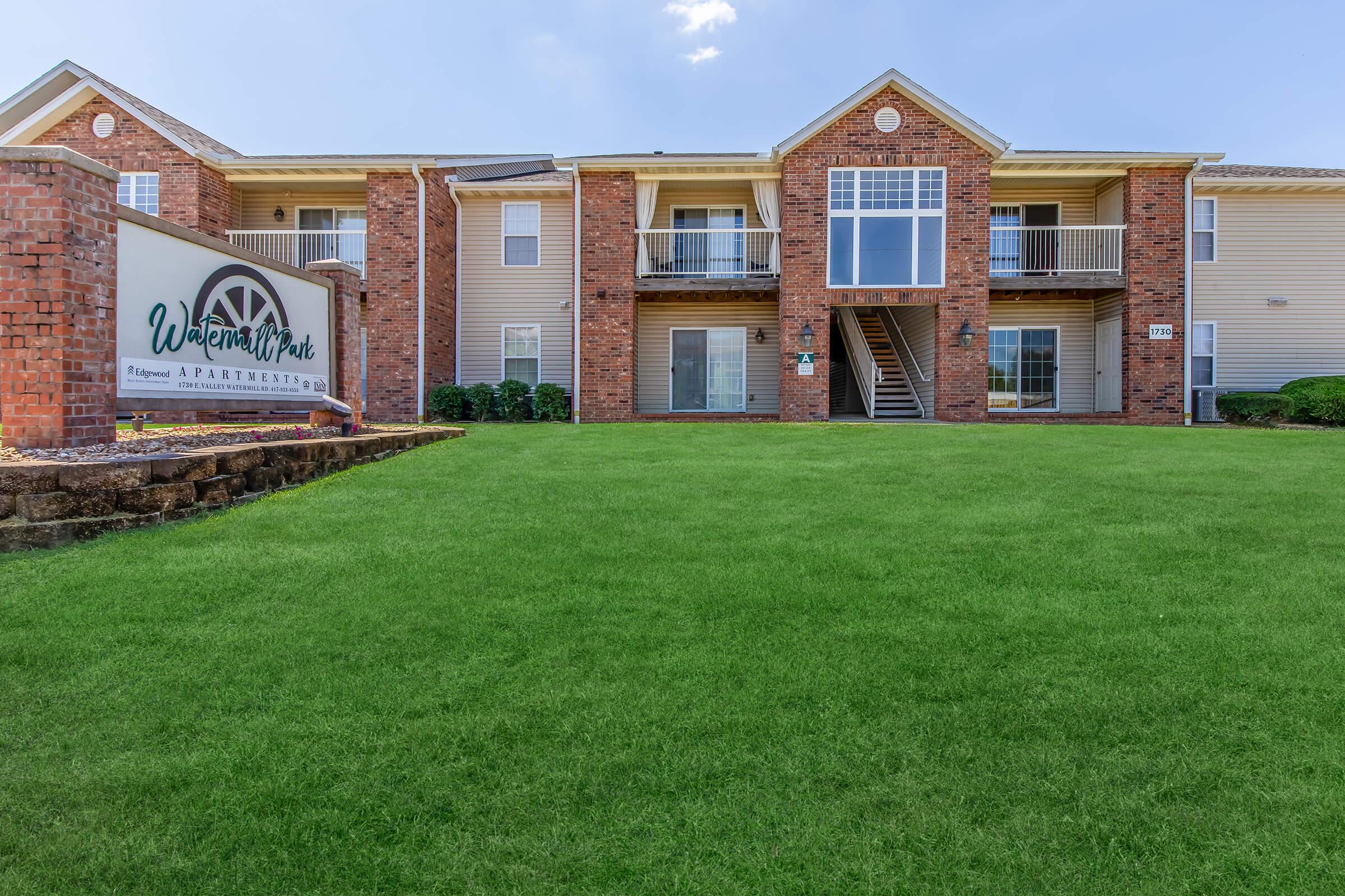 Exterior view of Watermark Park Apartments featuring a well-maintained green lawn, a brick building with multiple balconies, and a stone sign at the front. The sky is clear with a few clouds, creating a bright and inviting atmosphere.