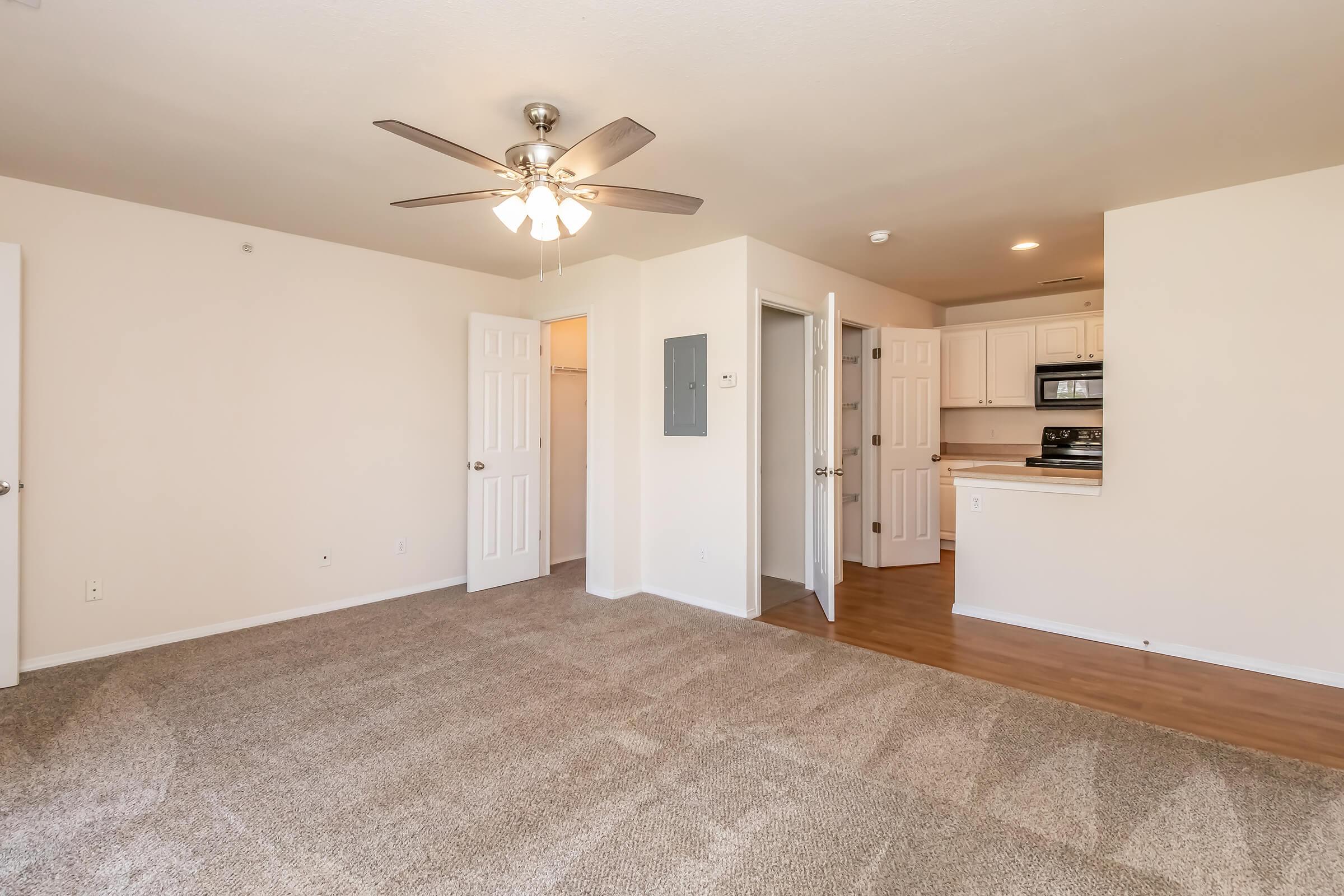 Spacious living area with light-colored walls and carpeted flooring. A ceiling fan is visible, along with an open kitchen area featuring white cabinetry and appliances. Doorways lead to other rooms, creating a cozy and inviting atmosphere. Natural light brightens the space.