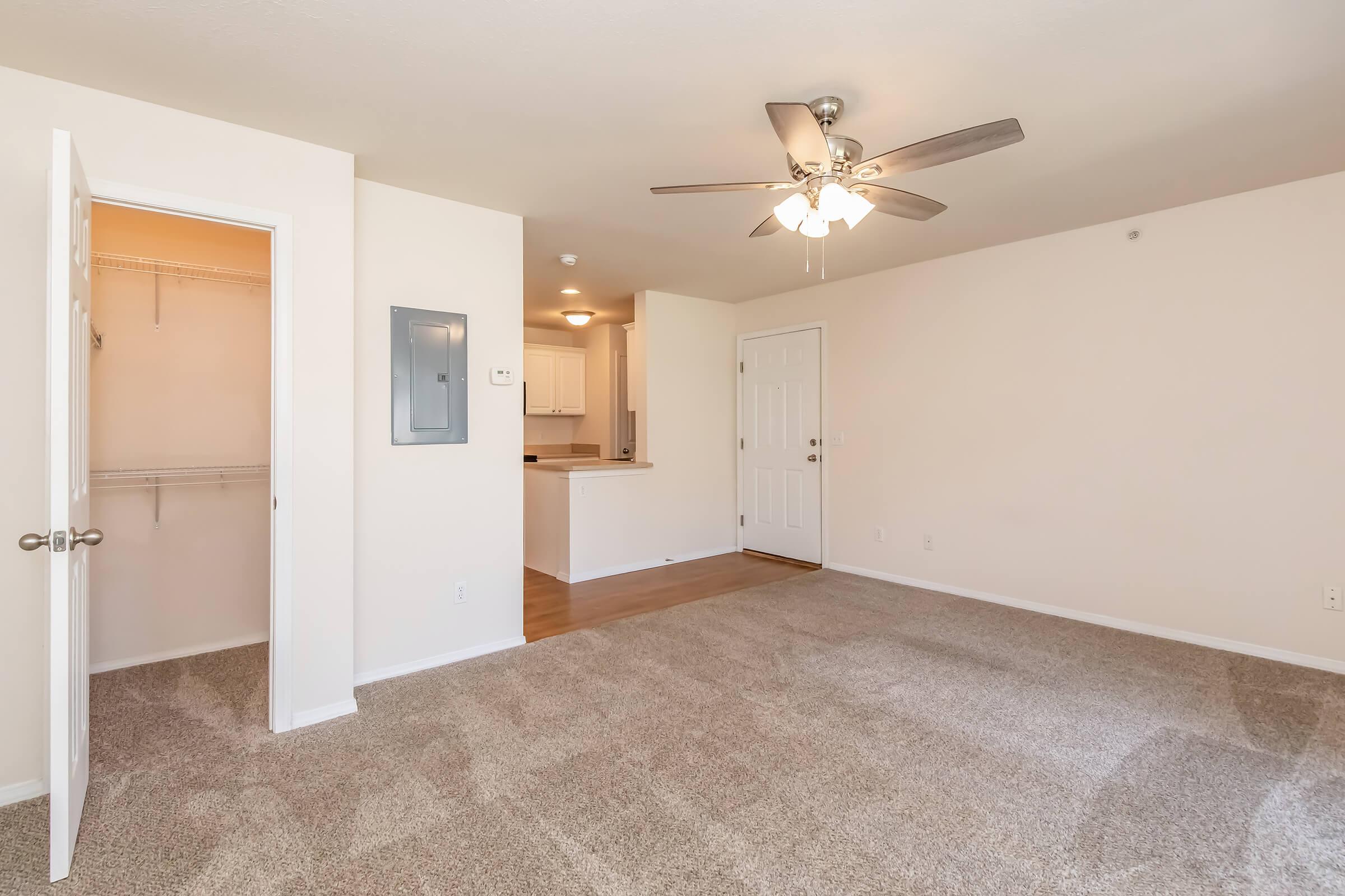 A spacious, well-lit living area featuring a ceiling fan, beige walls, and light brown carpet. An open doorway leads to a closet, and another doorway opens to a kitchen area with white cabinetry. The room has a welcoming atmosphere, inviting natural light through the entrance.