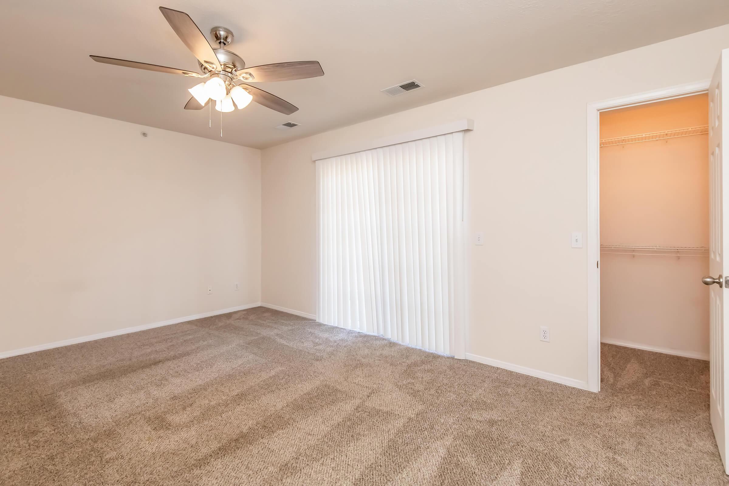 A spacious room with light beige walls and carpet flooring. The ceiling features a ceiling fan with lights. There is a large sliding glass door with sheer vertical blinds that leads outside, and a doorway to a closet on the right. The room is well-lit and appears to have neutral decor.