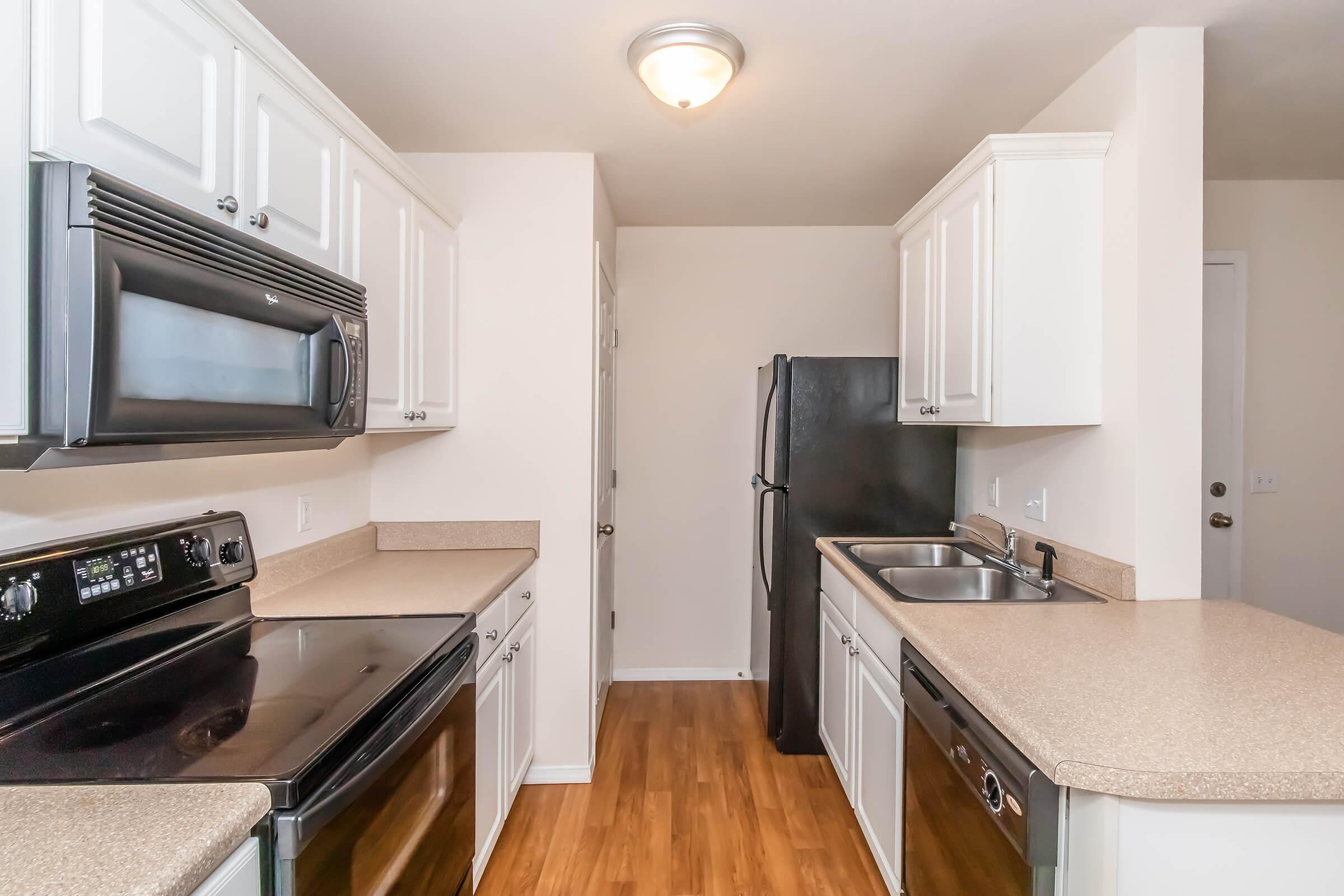 A modern kitchen featuring white cabinets, black appliances, and light countertops. The layout includes a refrigerator, an oven, and a sink, with wood-style flooring and neutral-colored walls creating a bright and inviting atmosphere.
