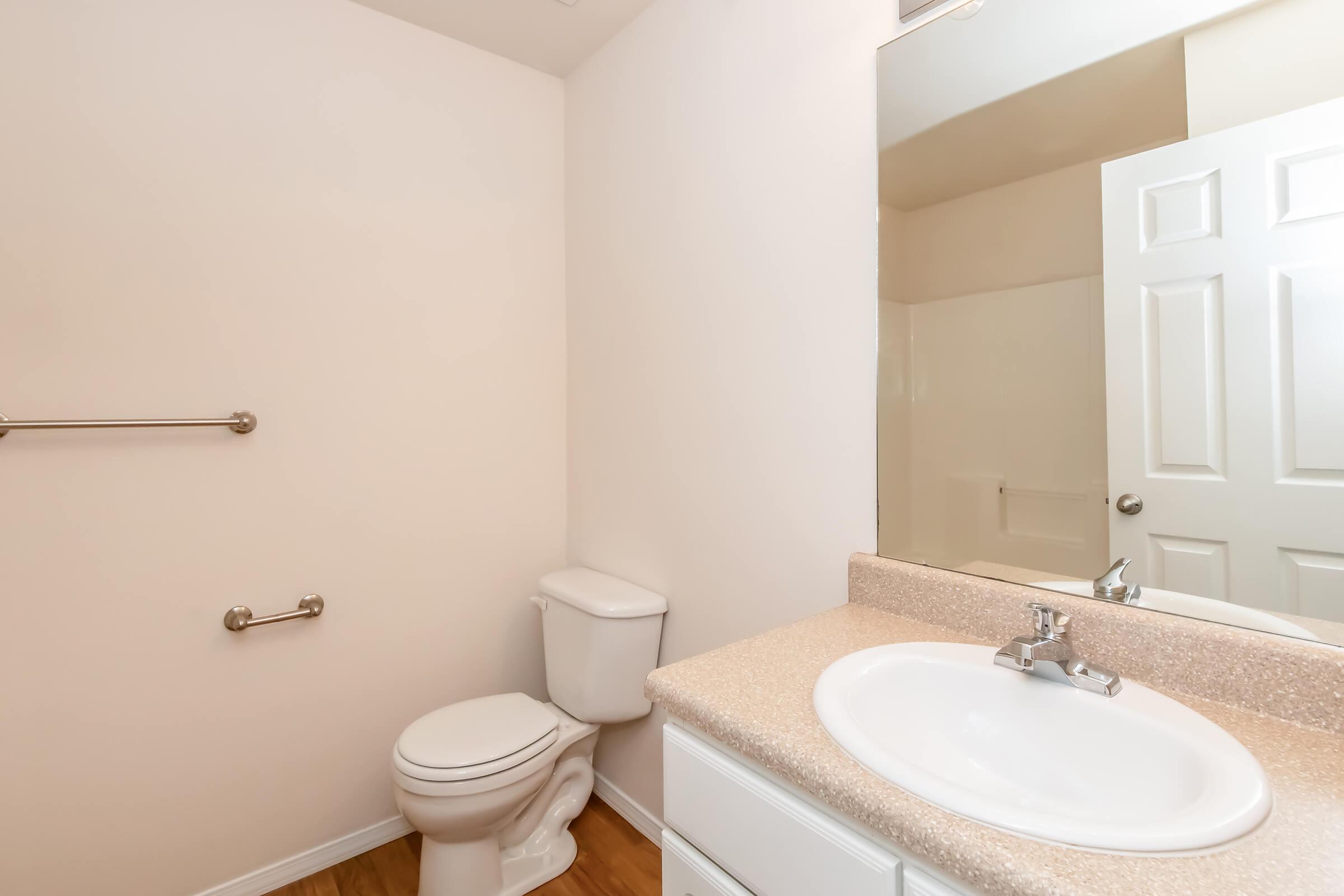 A clean and modern bathroom featuring a toilet, a sink with a countertop, a mirror above the sink, and a towel bar on the wall. The walls are painted in a light color, and the floor has a wooden finish. A white door is visible, leading to another area.