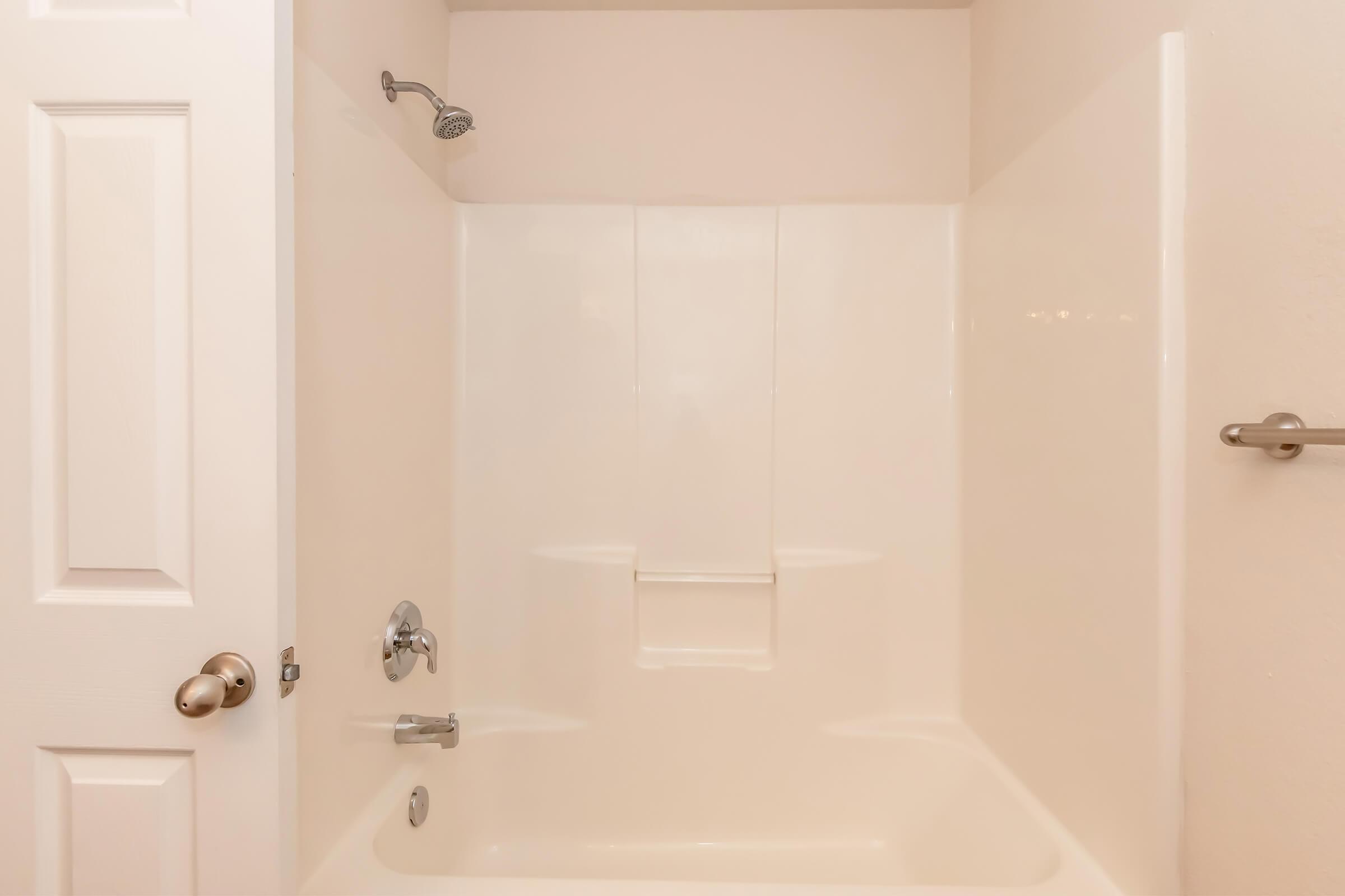 A clean, empty bathroom with a white shower and tub. The walls are smooth and light-colored, featuring a showerhead and a grab bar. A closed door with a round doorknob is visible on the left side. The space appears bright and well-maintained, ideal for a simple bathing setup.
