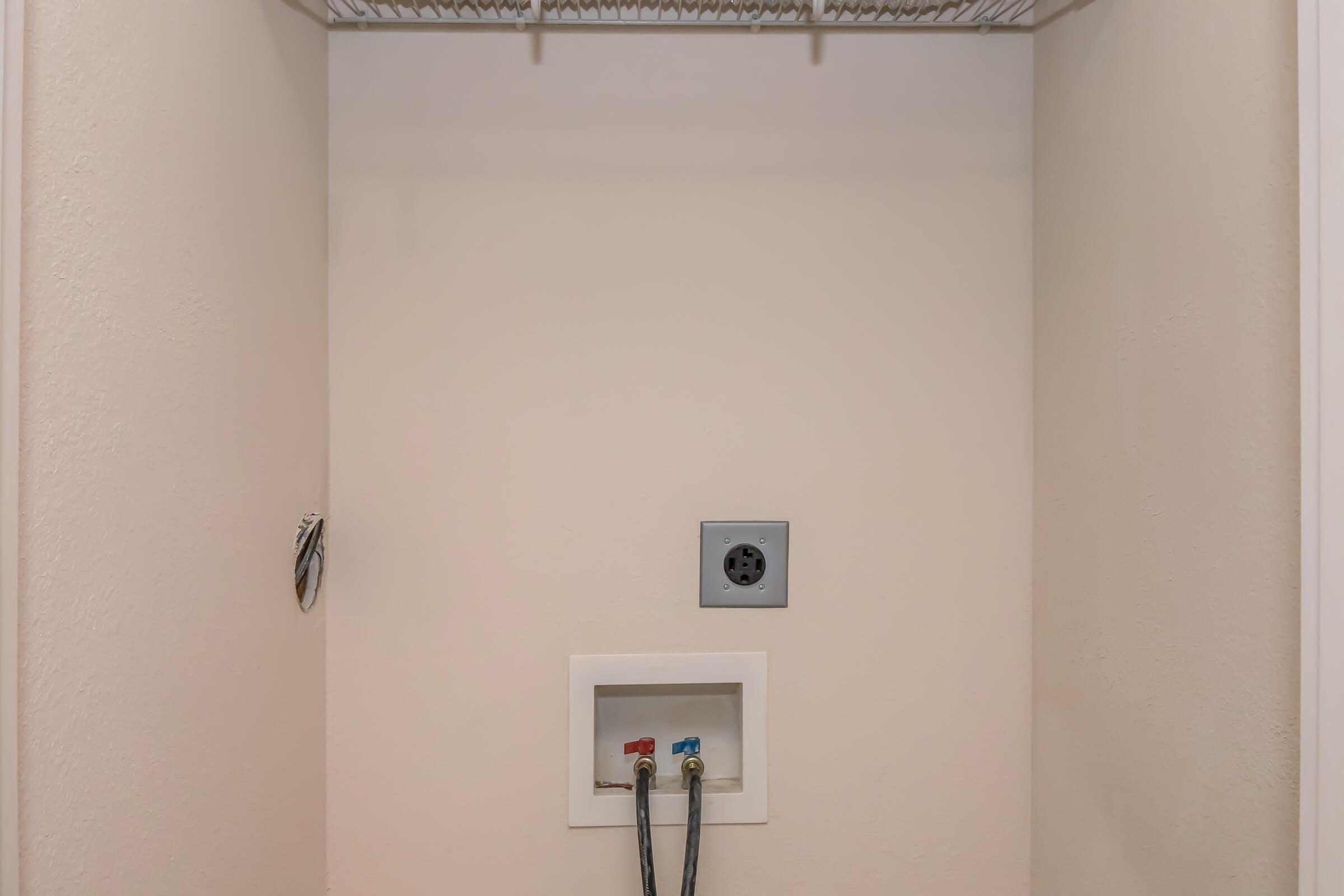 Empty wall space with utility connections. Two colored pipes (red and blue) for hot and cold water are visible at the bottom, along with an electric outlet in a rectangular box. The wall has a smooth texture and is painted in a neutral color, with a wire shelf above, indicating potential for storage.