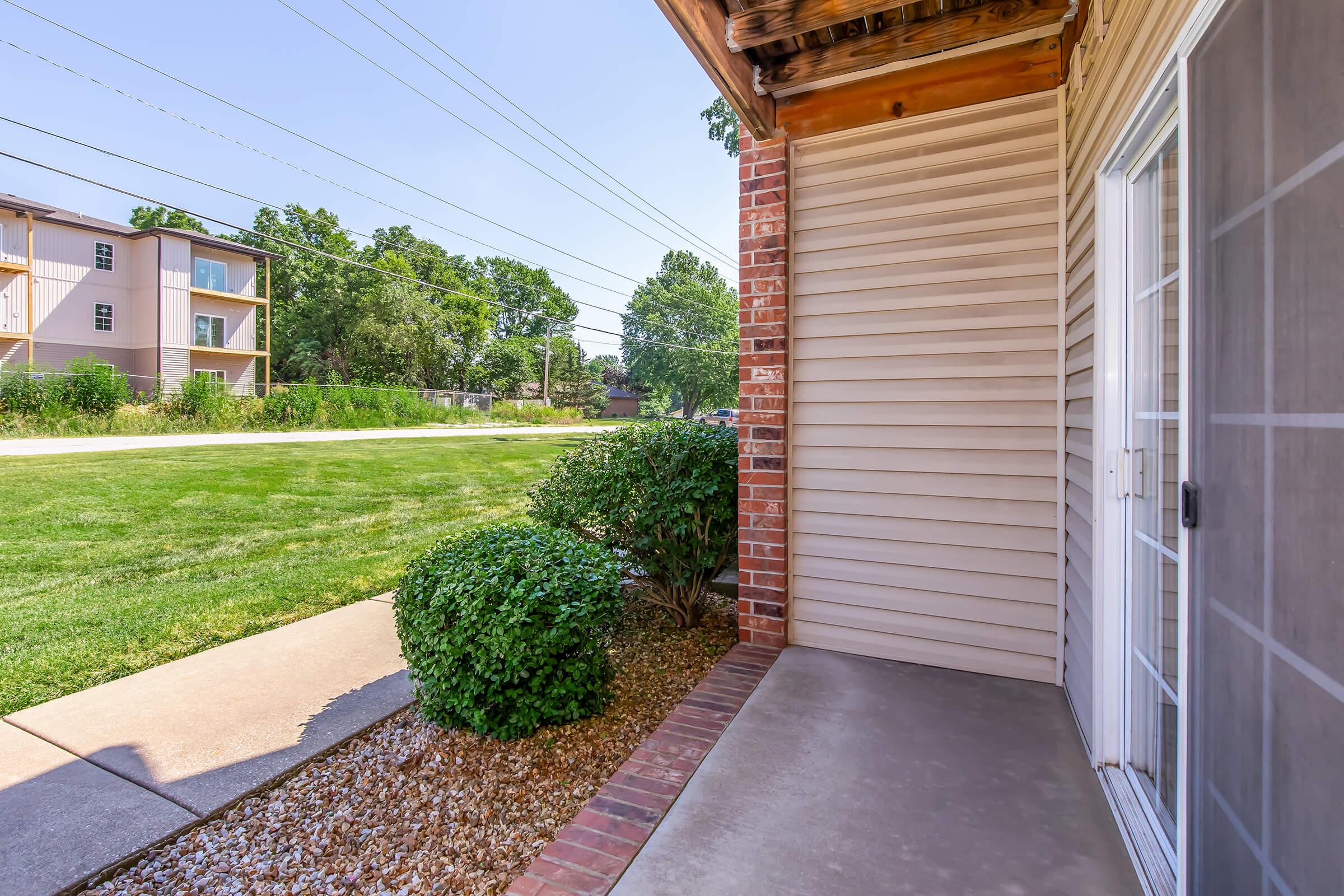 A view of a front porch area with a concrete pathway leading to a grassy yard. There are bushes along the pathway, and a nearby building is visible in the background. The sky is clear and blue, indicating a sunny day. Power lines are visible overhead.