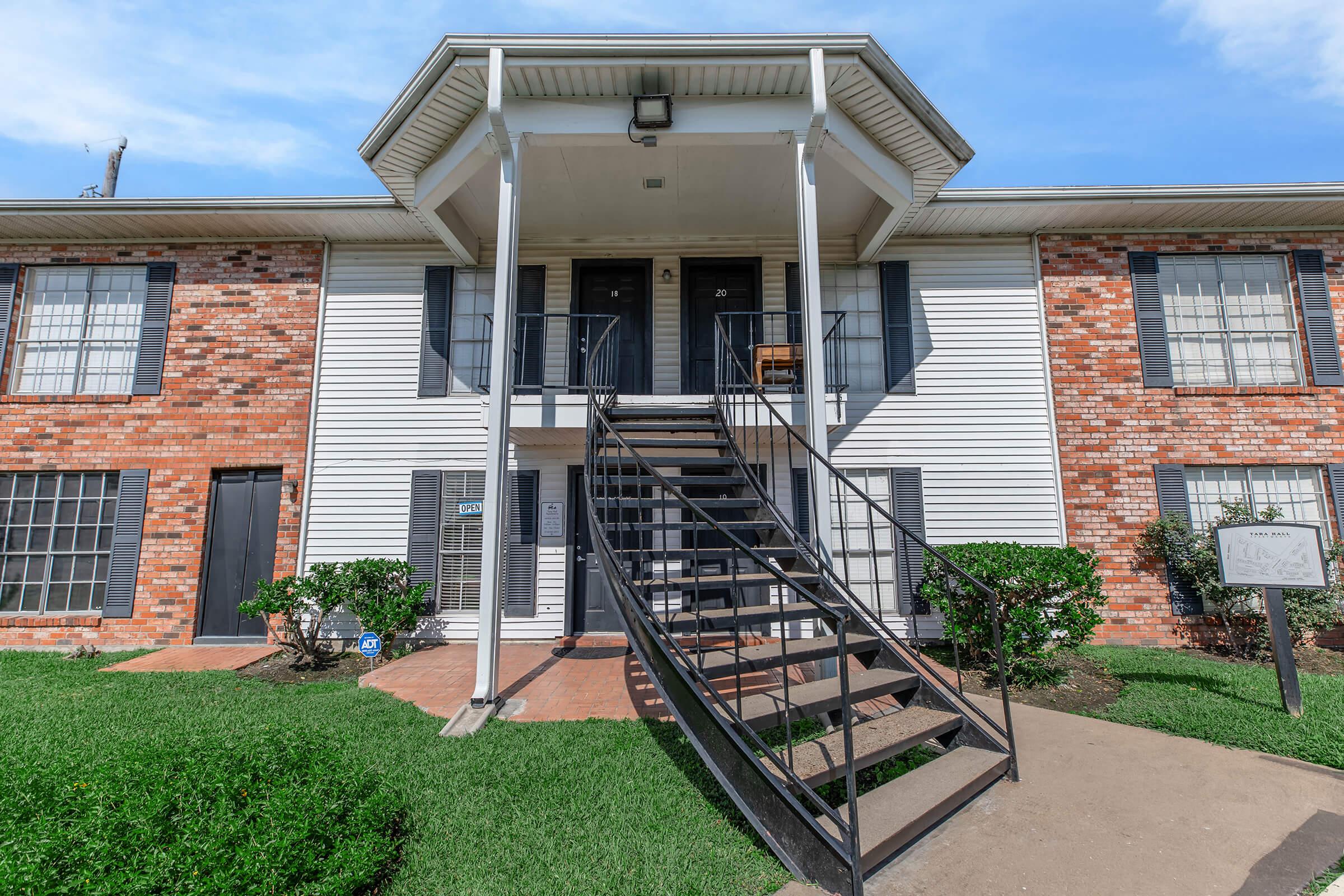 Exterior view of a two-story apartment building featuring a brick facade. A black metal staircase leads to the upper level, with a small landing and door visible. Green grass and bushes are well-maintained around the entrance, under a clear blue sky.