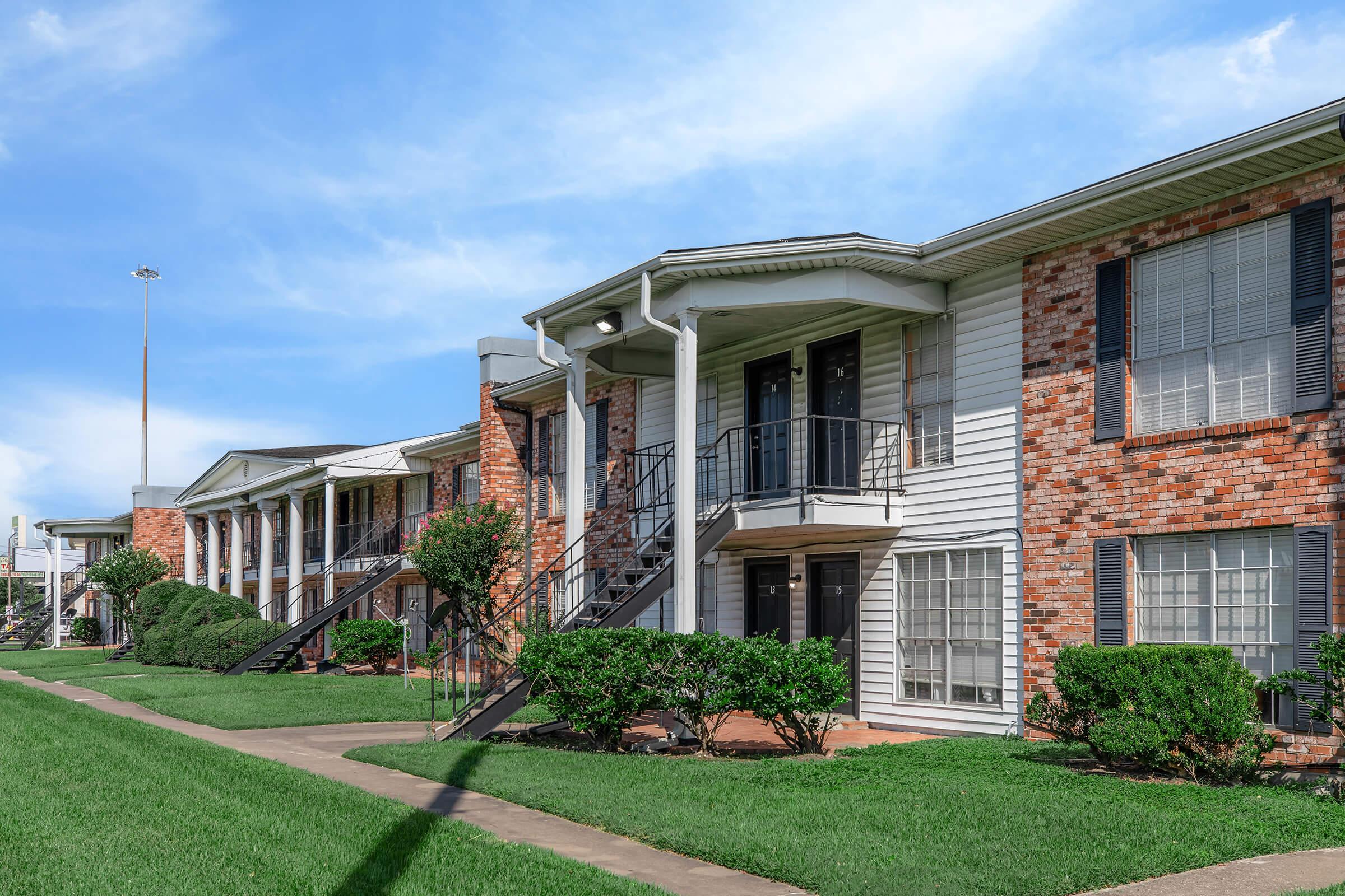 A row of two-story apartment buildings with a mix of brick and siding exteriors. Each building features a staircase leading to the upper level. Well-maintained grassy areas and shrubs line the sidewalks, and the sky is clear with a few clouds.