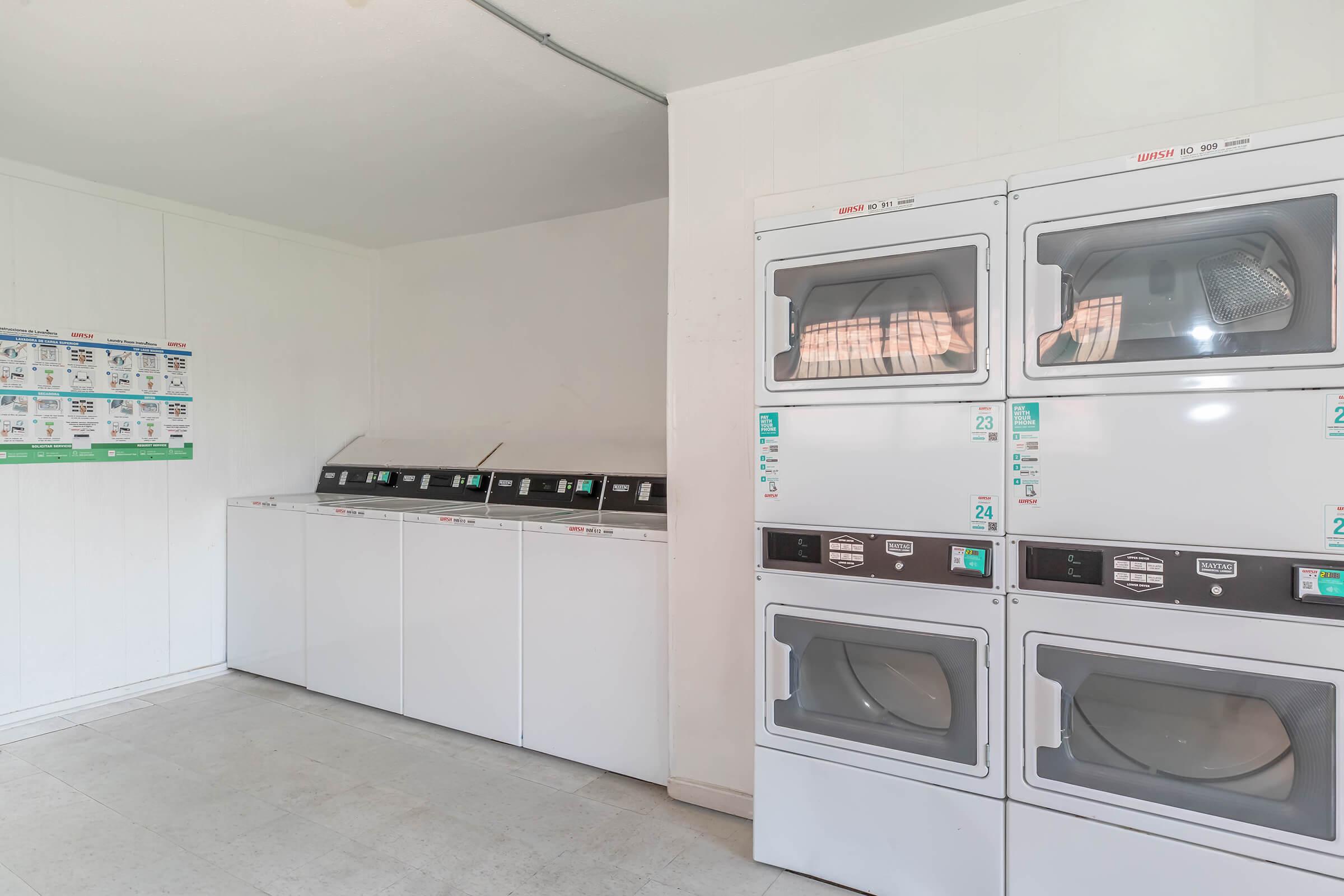 A clean laundromat interior featuring two rows of white washing machines and dryers. The machines are organized against a wall, with signs and instructions displayed nearby. The space is bright and tidy, with tiled flooring and a simple, modern design.